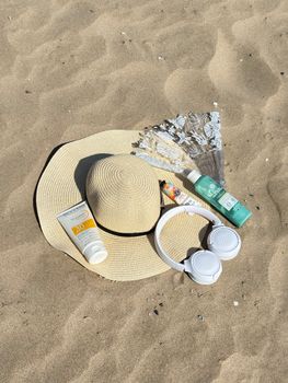 Flat lay of beach essentials including sun hat, sunscreen, headphones, and fan on sandy beach.