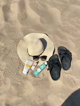 Flat lay of beach essentials including sunglasses, sunscreen, hat, and sandals on sandy beach.