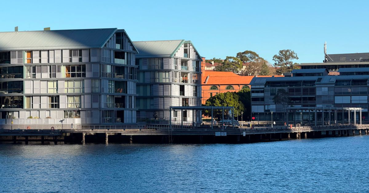 Modern waterfront buildings with glass facades under a clear blue sky, reflecting on the water.