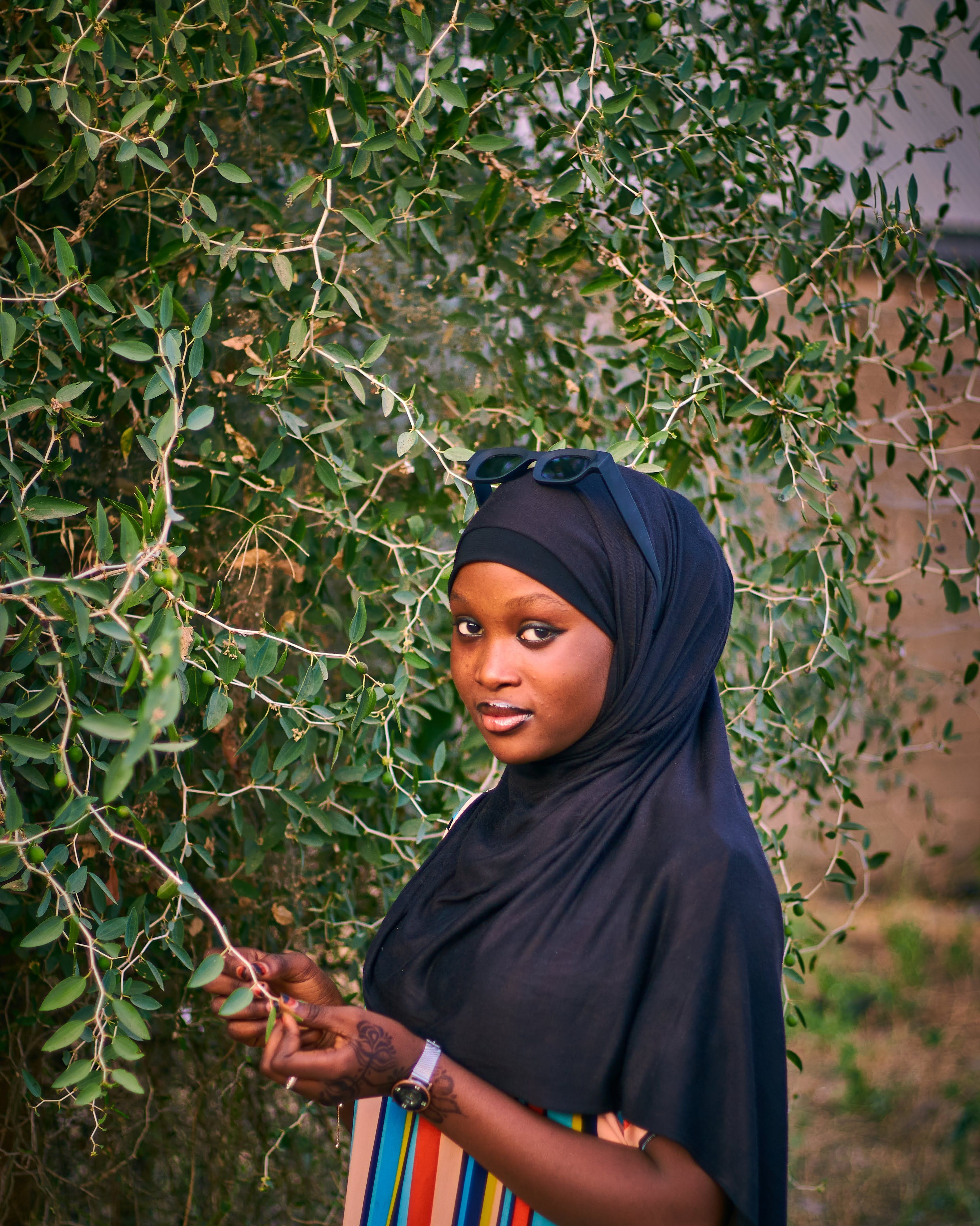 A young woman in a hijab stands beside a vibrant green tree branch outdoors.