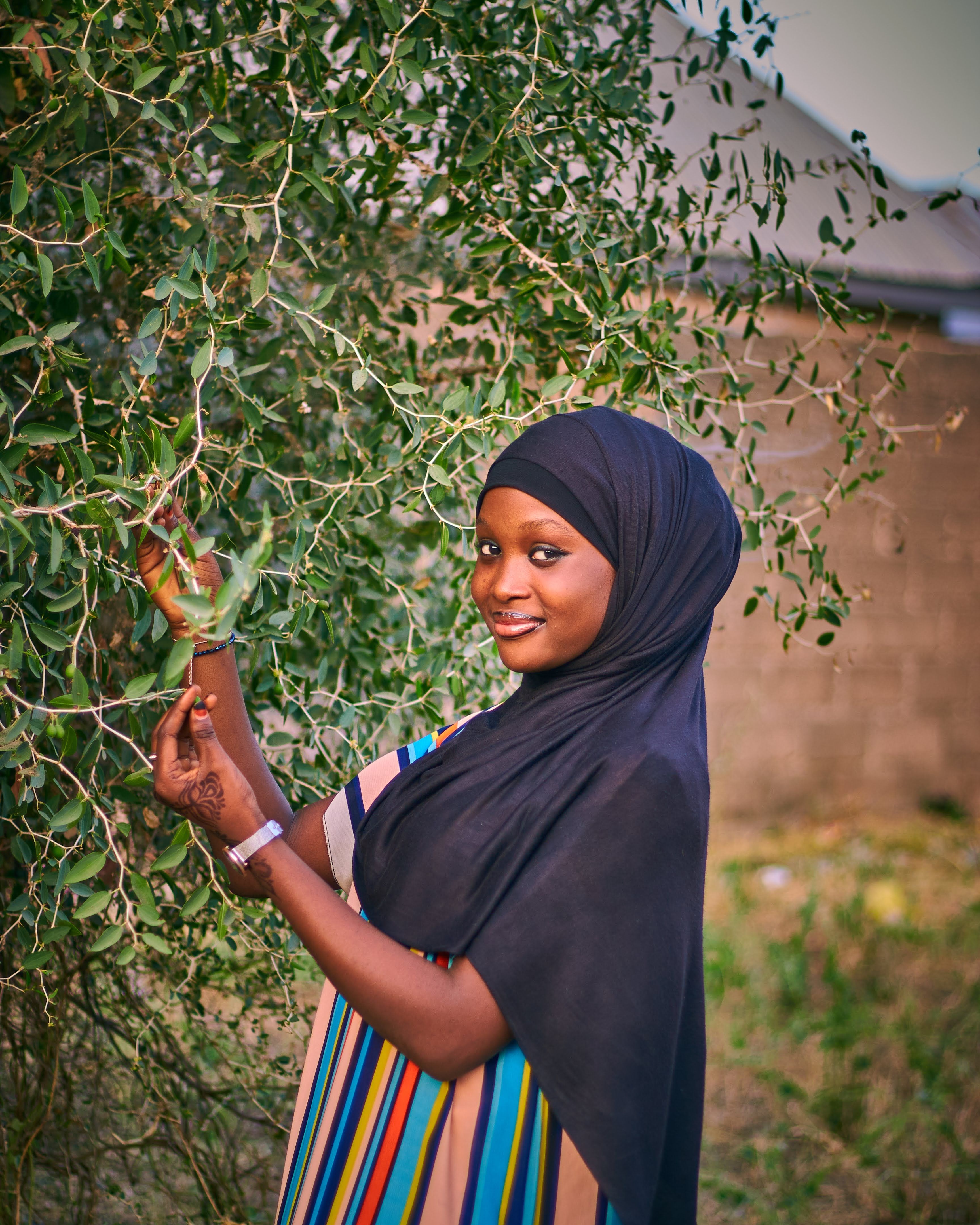 A woman wearing a hijab smiles while standing next to a tree, holding a smartphone.