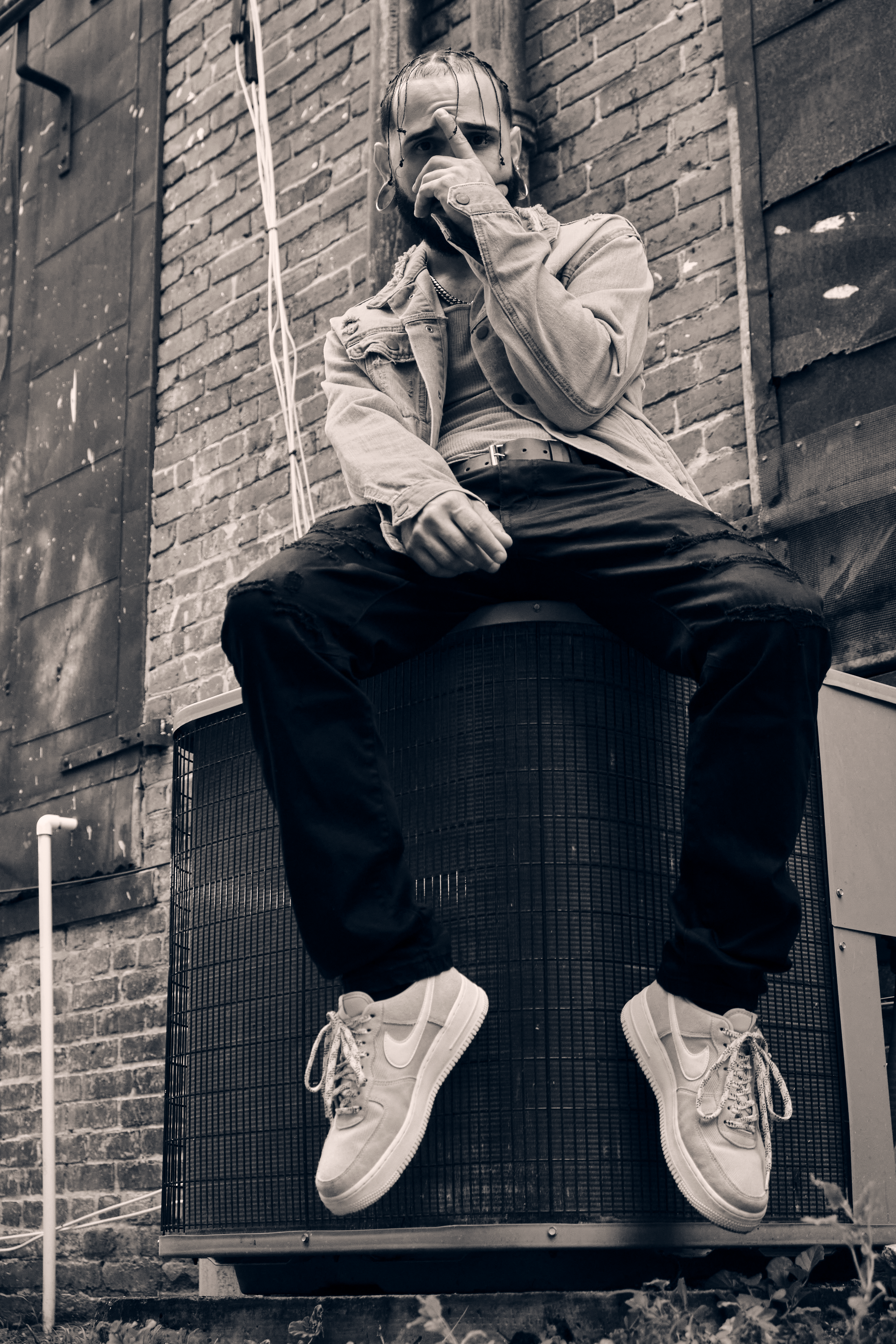 Stylish man sitting on AC unit against a gritty brick wall outdoors.