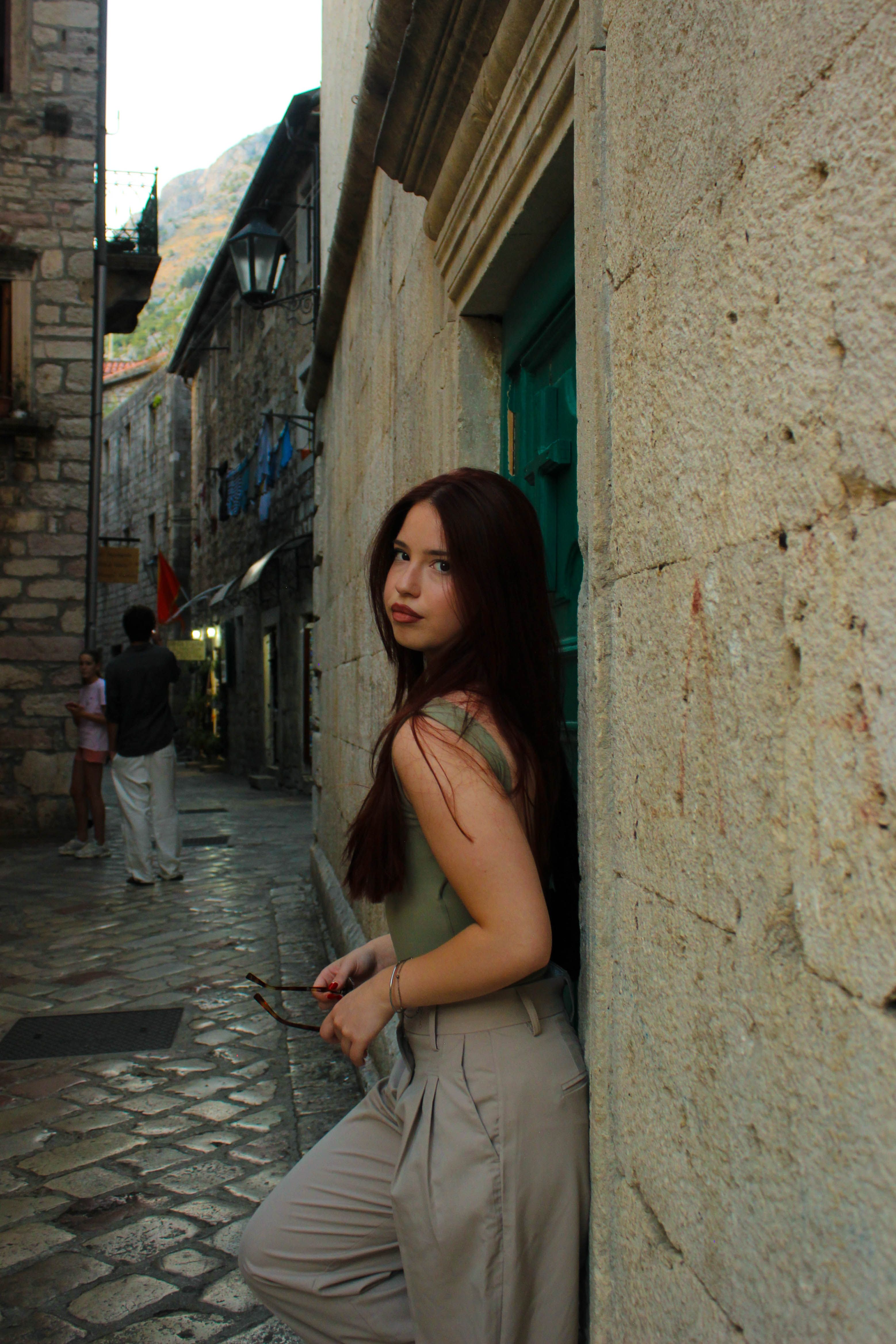 Stylish young woman with red hair leans against a wall in a historic European alleyway.