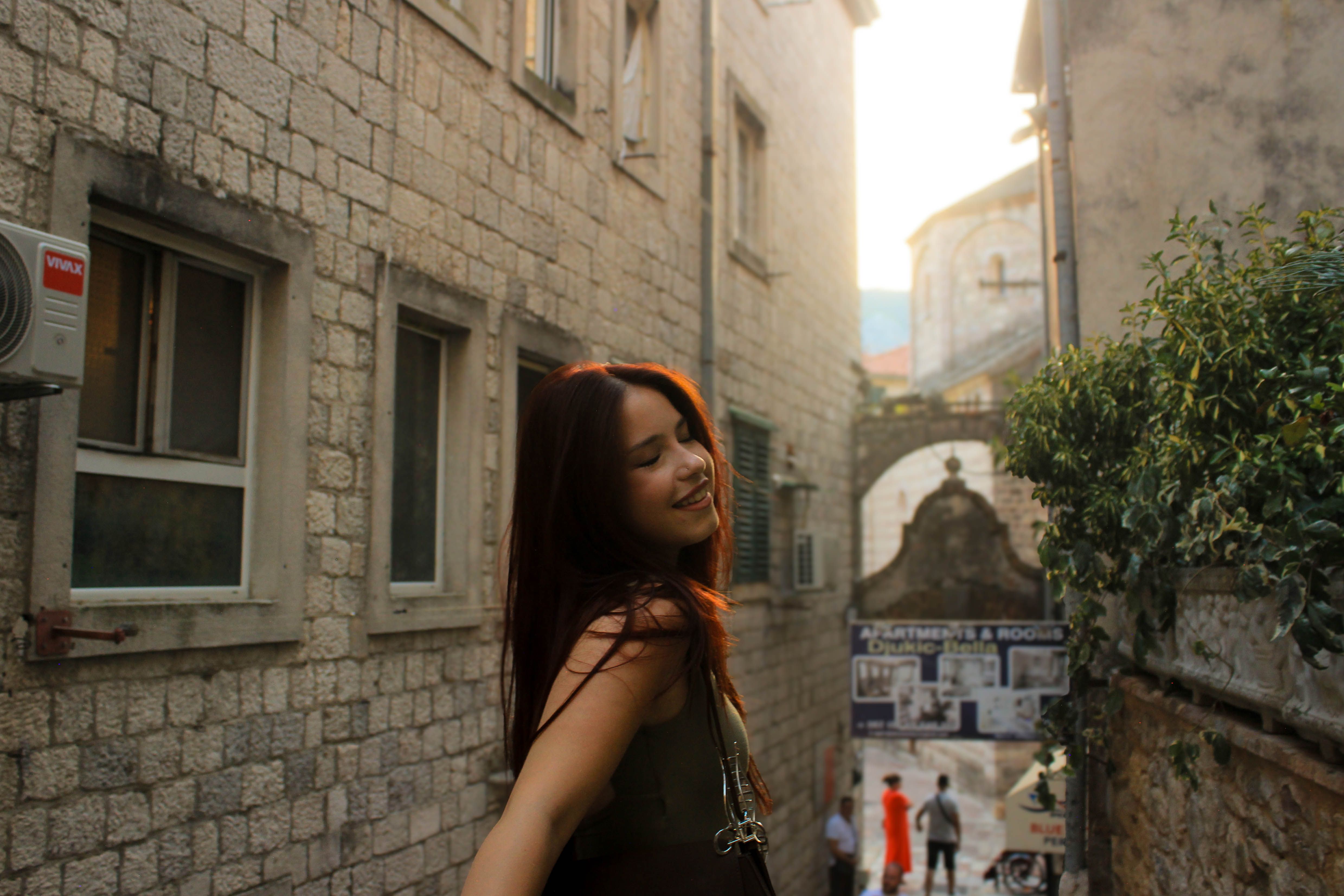 A young woman joyfully smiles while exploring a historic stone alleyway in a sunlit European street.