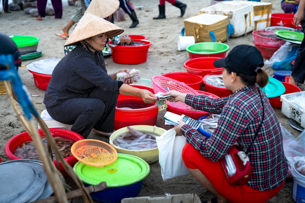 Asian female in traditional authentic Vietnamese hat sitting on stool in street fish market and selling seafood to female customer