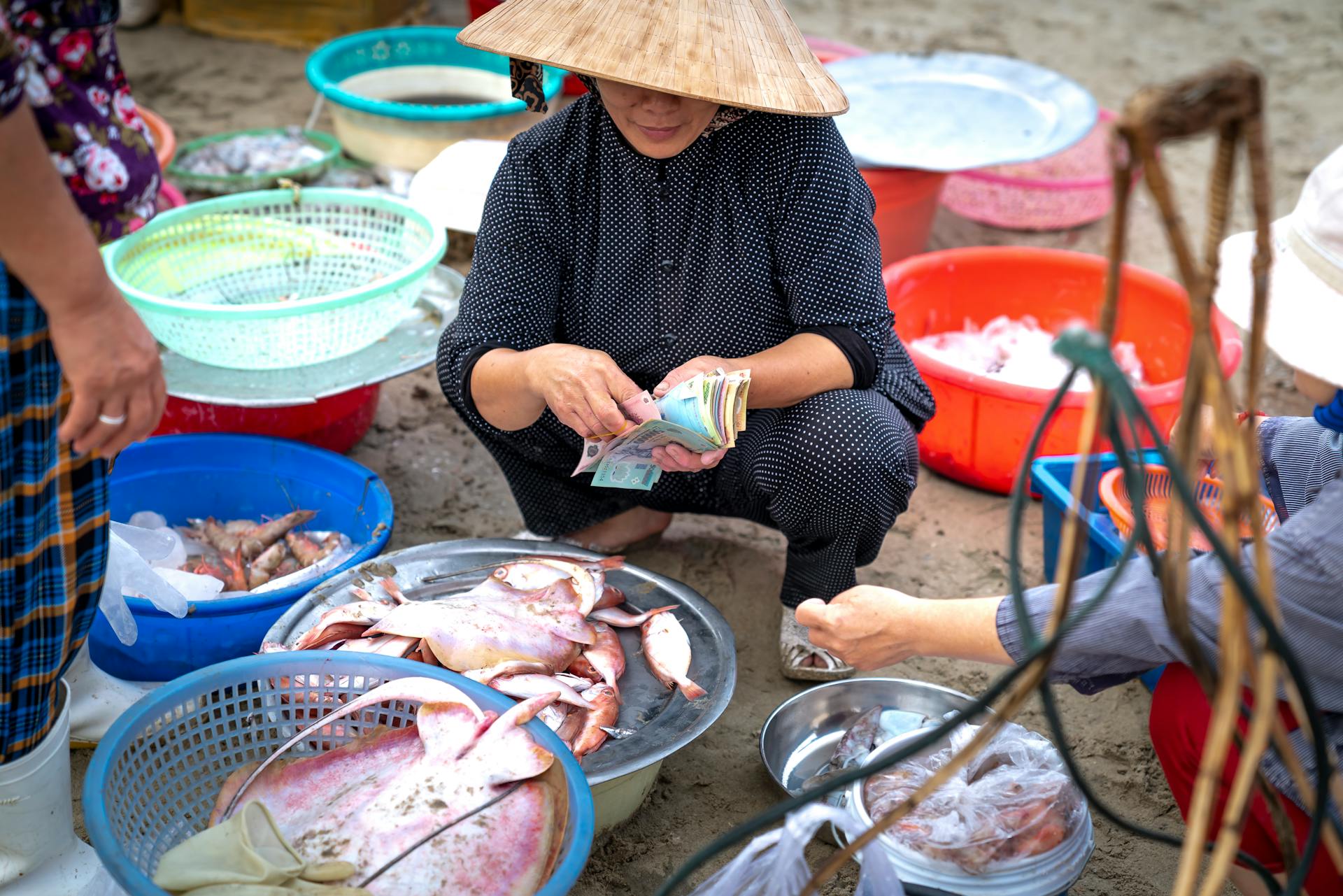 A vendor counting money in an Asian seafood market while surrounded by fresh catches and colorful baskets.