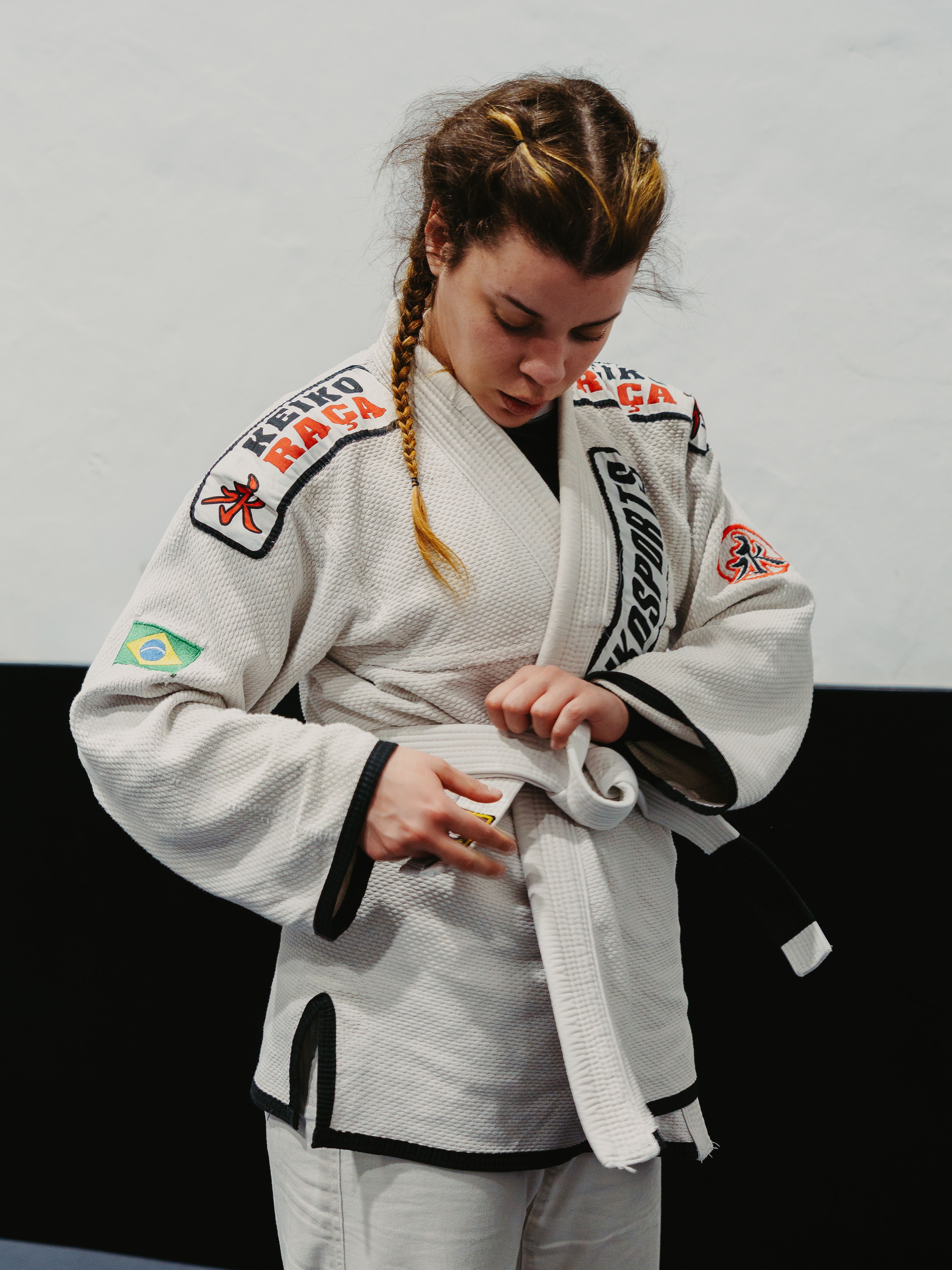 Young woman in Brazilian jiu-jitsu gi tying her belt in a dojo setting.