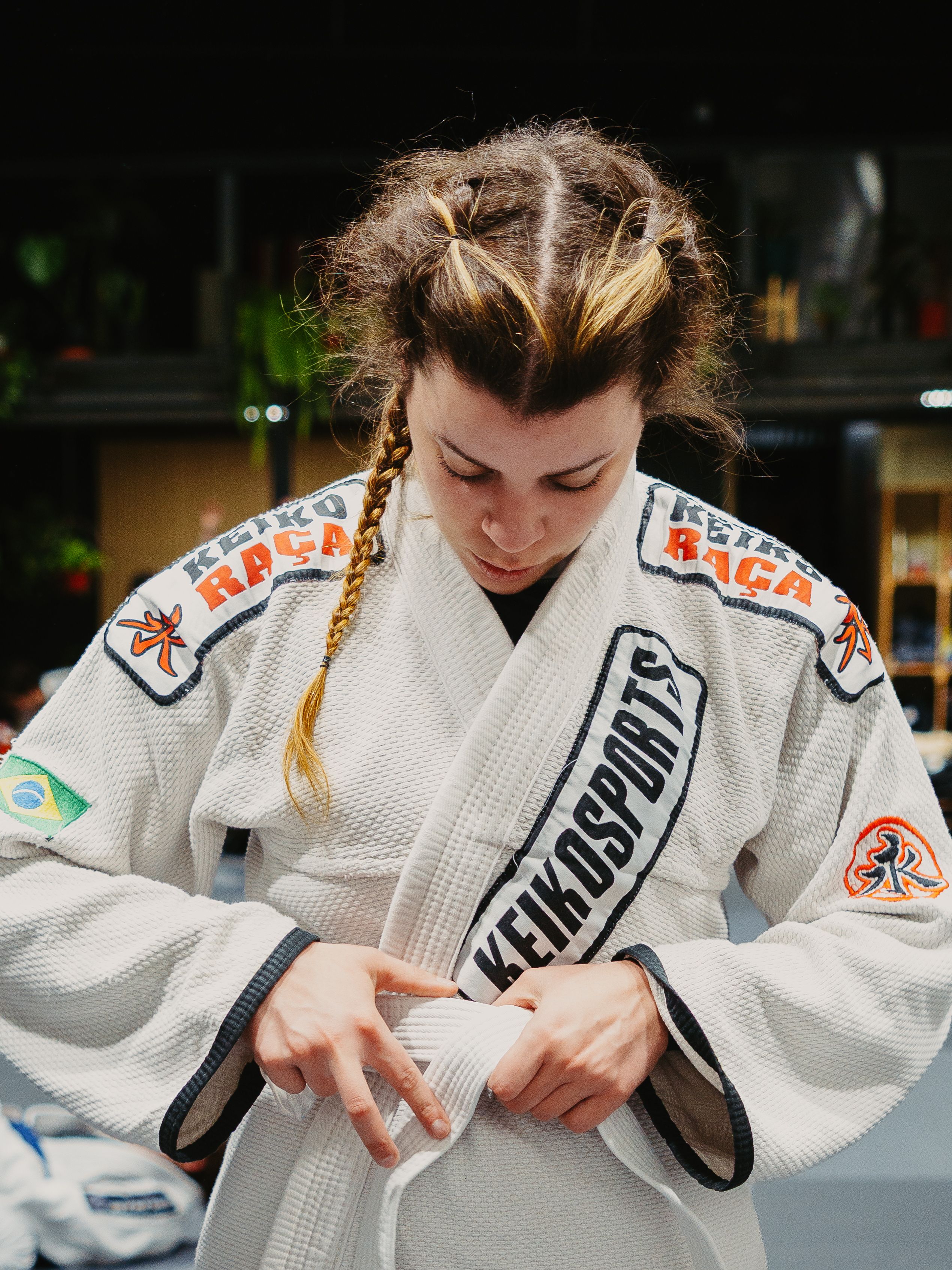 A focused female athlete adjusts her gi belt before Brazilian Jiu-Jitsu practice indoors.