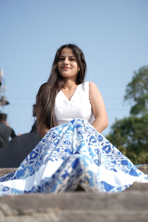 Free Smiling woman sitting on steps outdoors in a vibrant blue and white dress under a clear sky. Stock Photo
