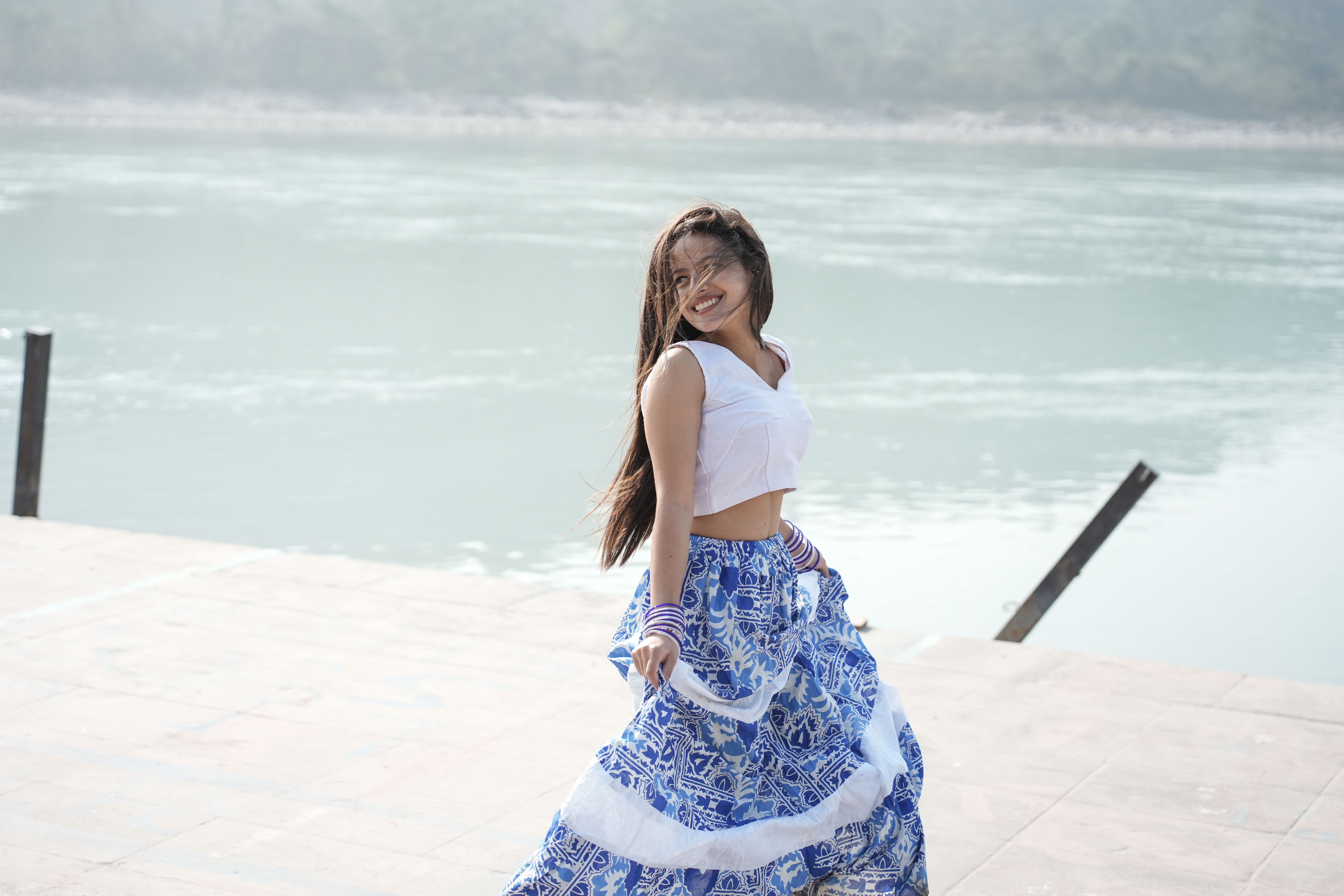 A woman in a white top and blue skirt dances joyfully by a calm river, exuding happiness.