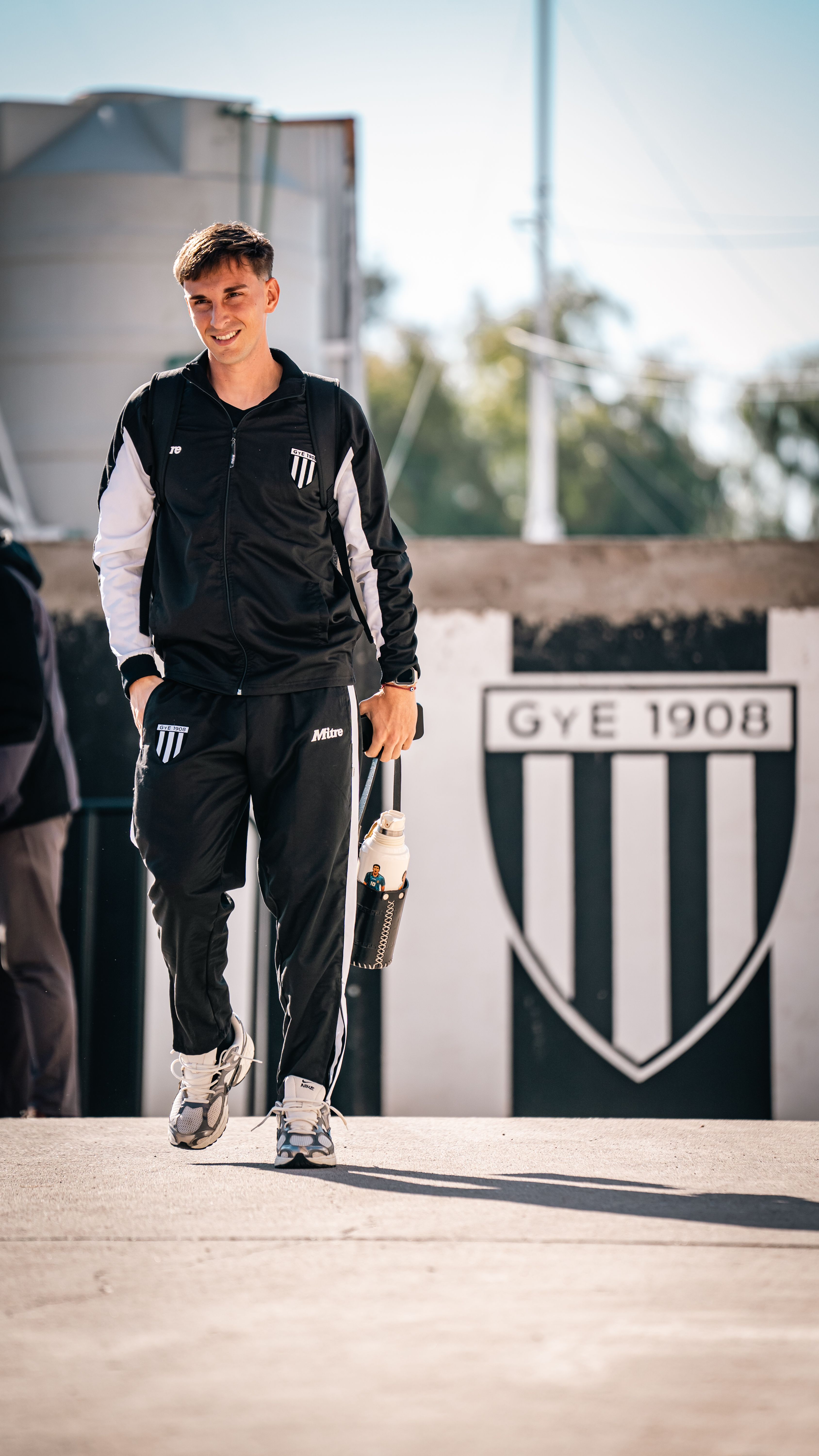 Young athlete smiling with sportswear beside GYE 1908 sports logo in sunny setting.