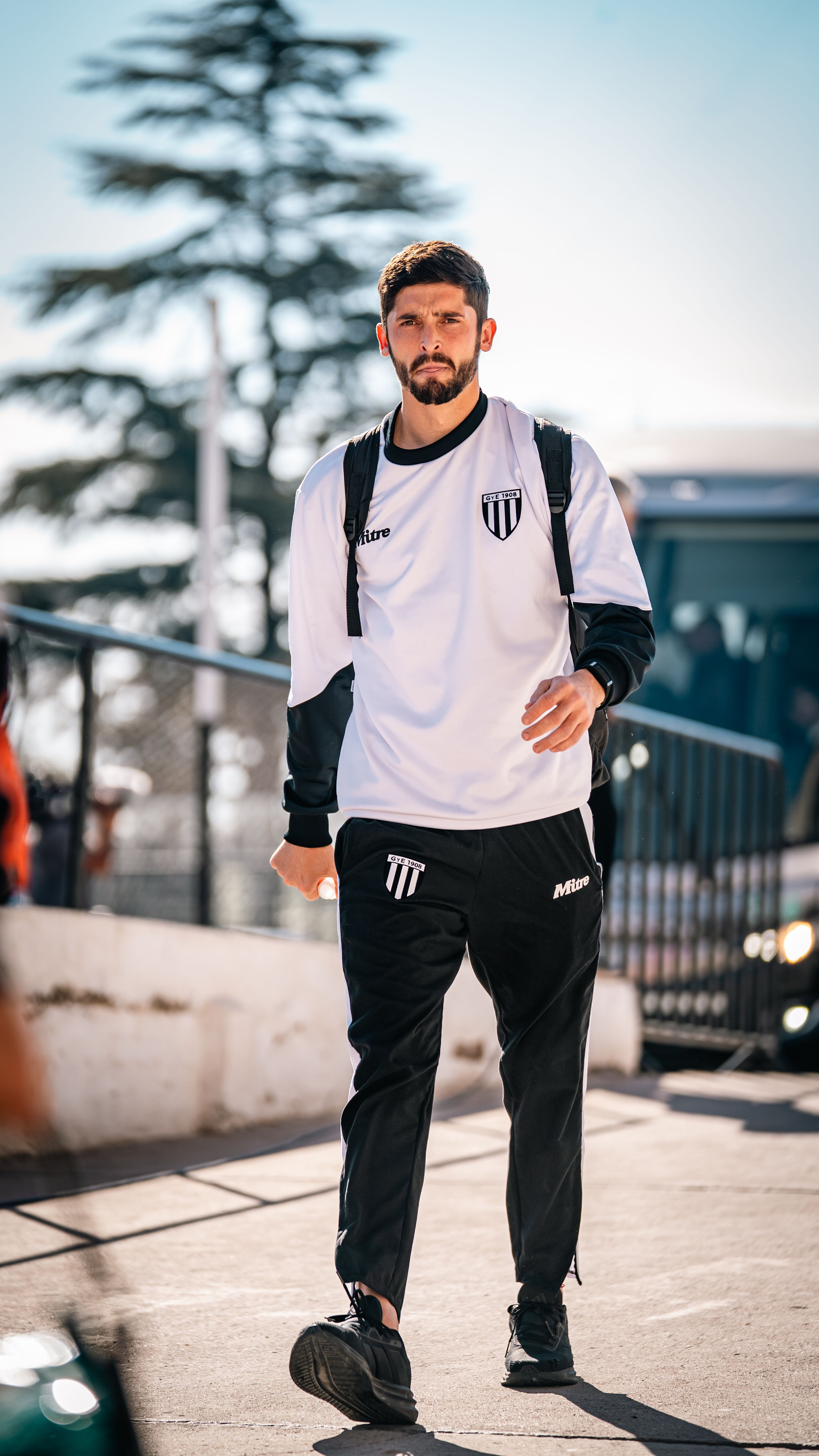 A male athlete wearing a white sports uniform and backpack walks outdoors in bright daylight.
