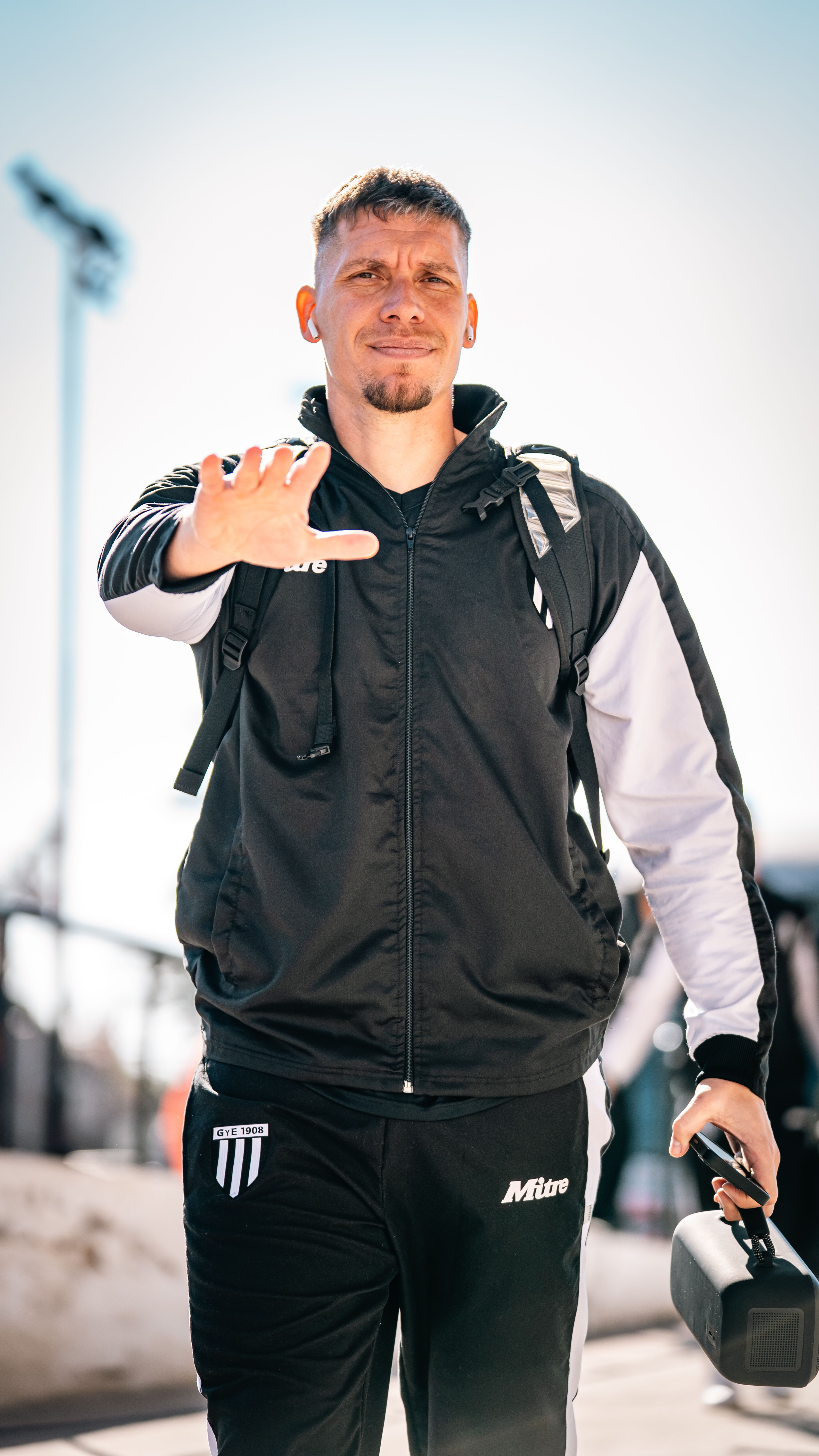 A sporty male adult walking outdoors holding headphones, dressed in casual athletic wear.