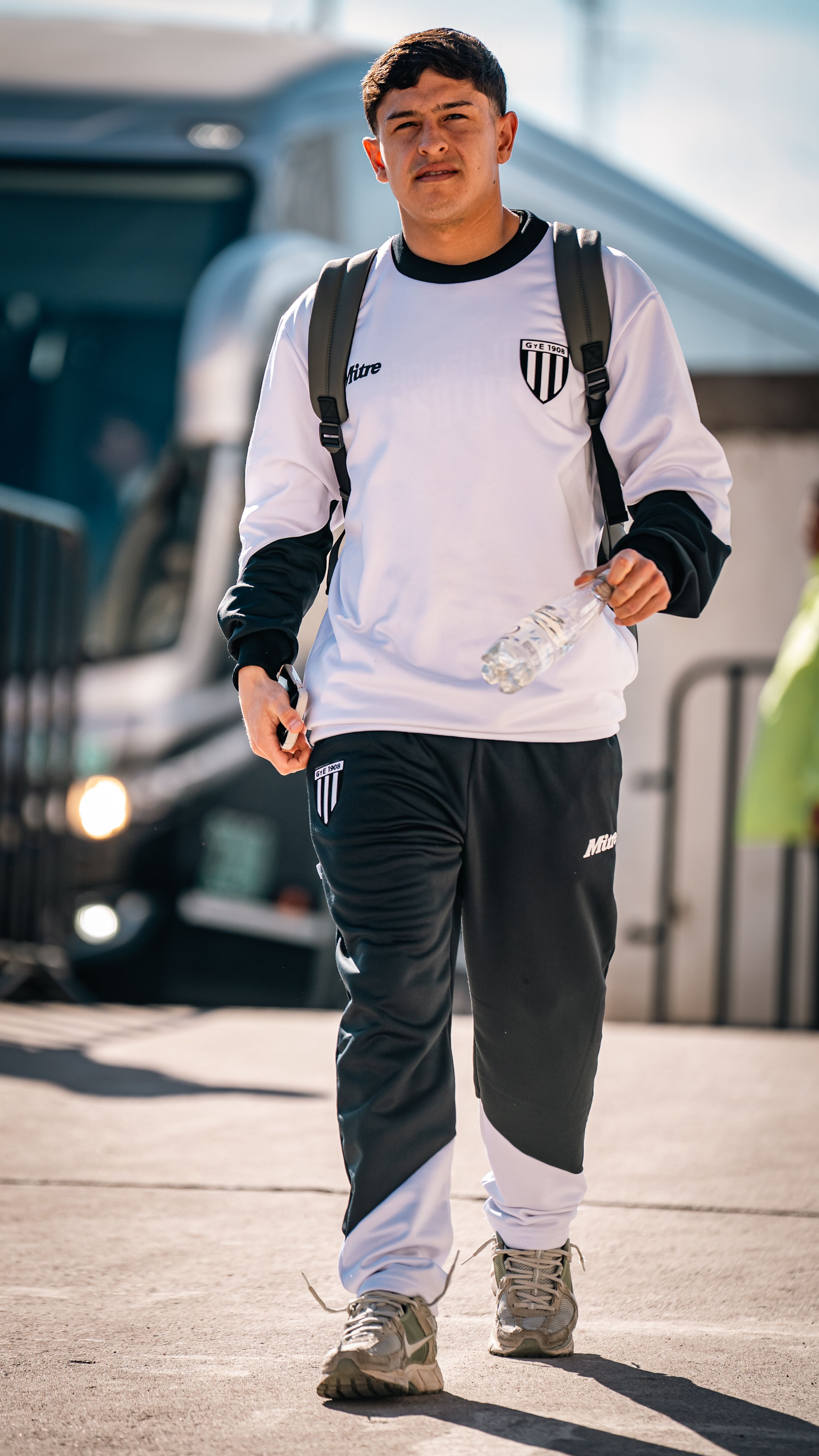 Young male athlete walks outdoors in team tracksuit, holding a water bottle. Bright, sunny day.