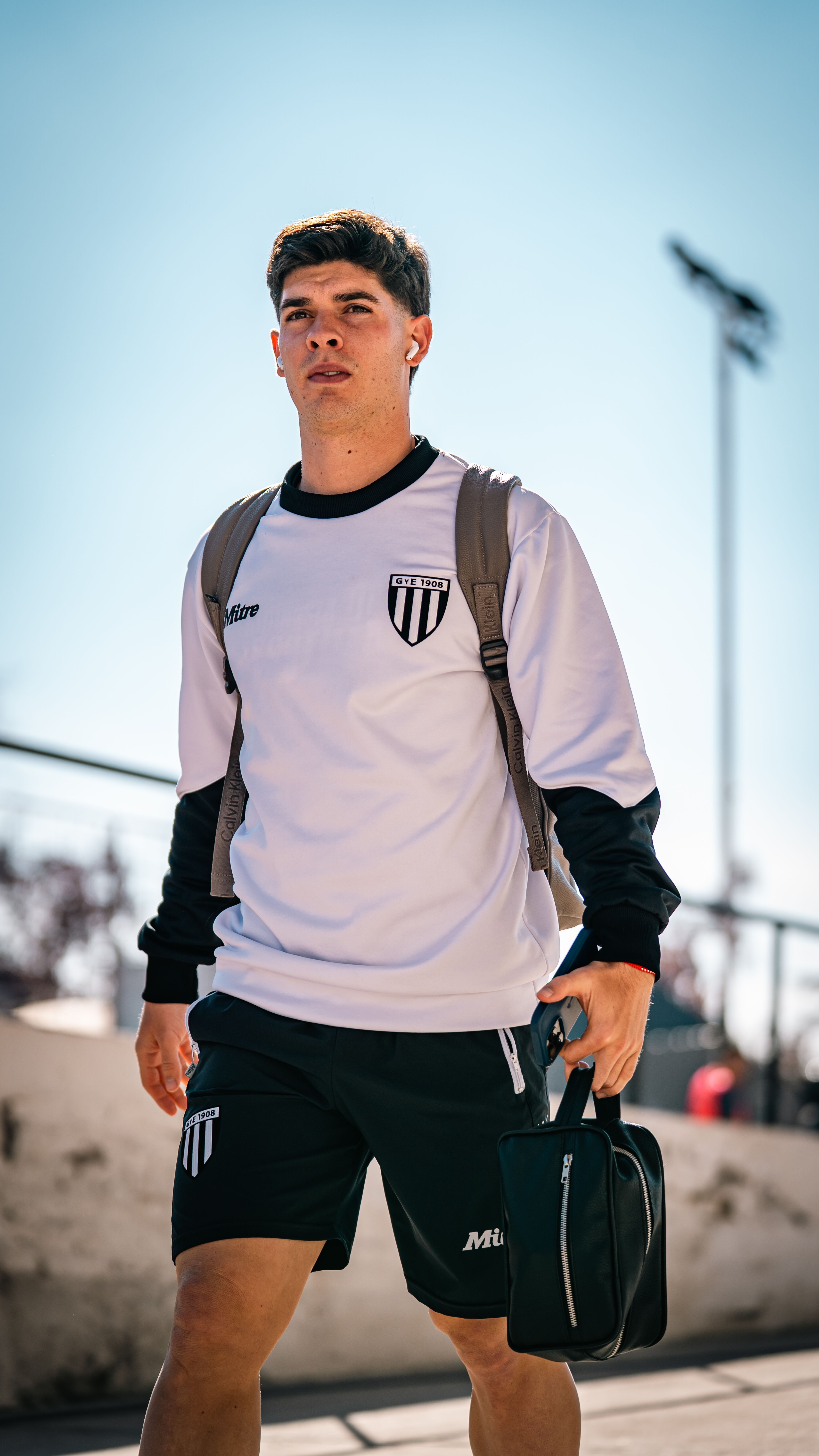 Young male athlete in sportswear walking outdoors on a sunny day, wearing a backpack and holding a bag.