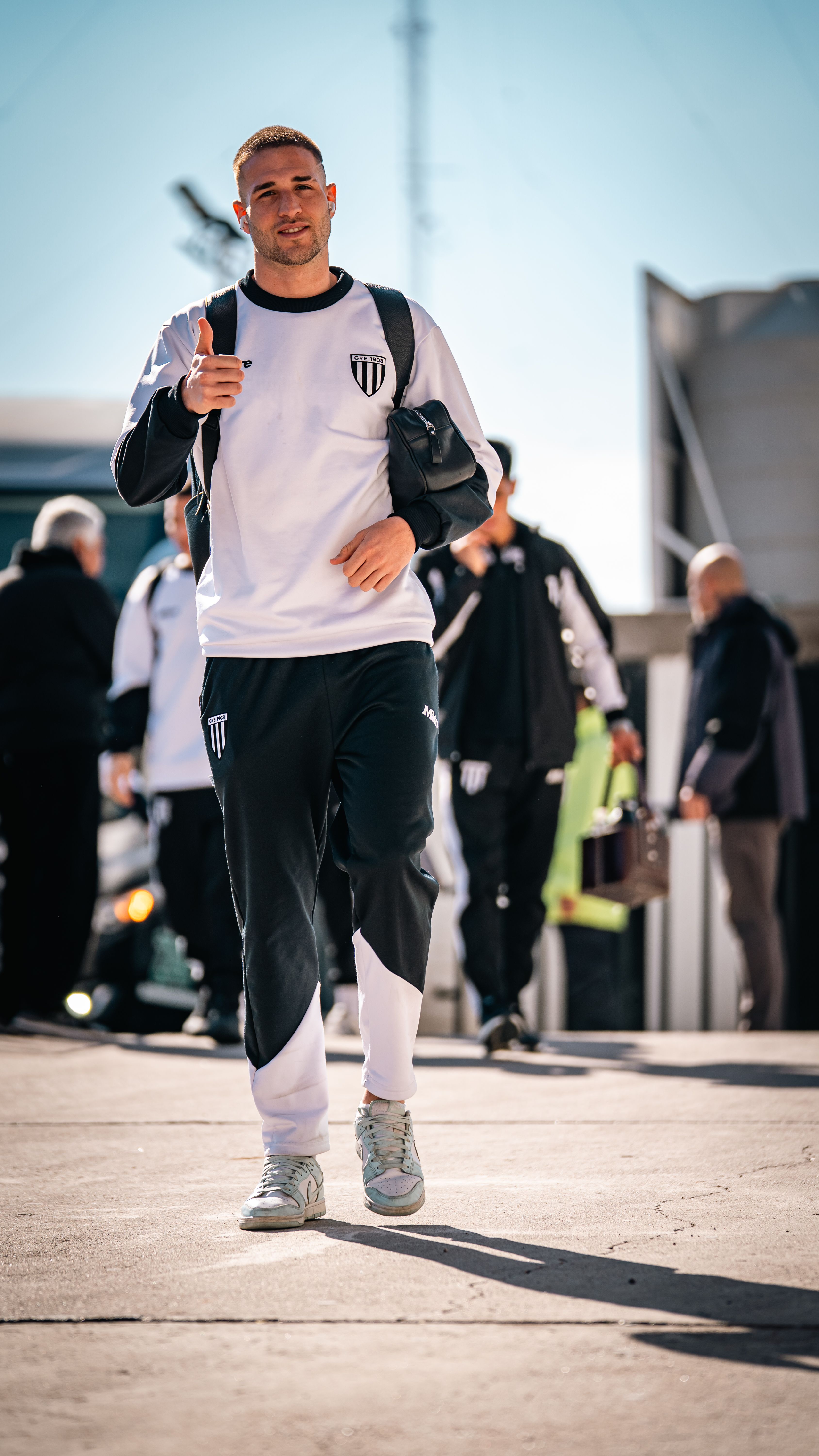 Sporty individual in athletic wear gives a thumbs-up gesture while walking on a sunny day outdoors.