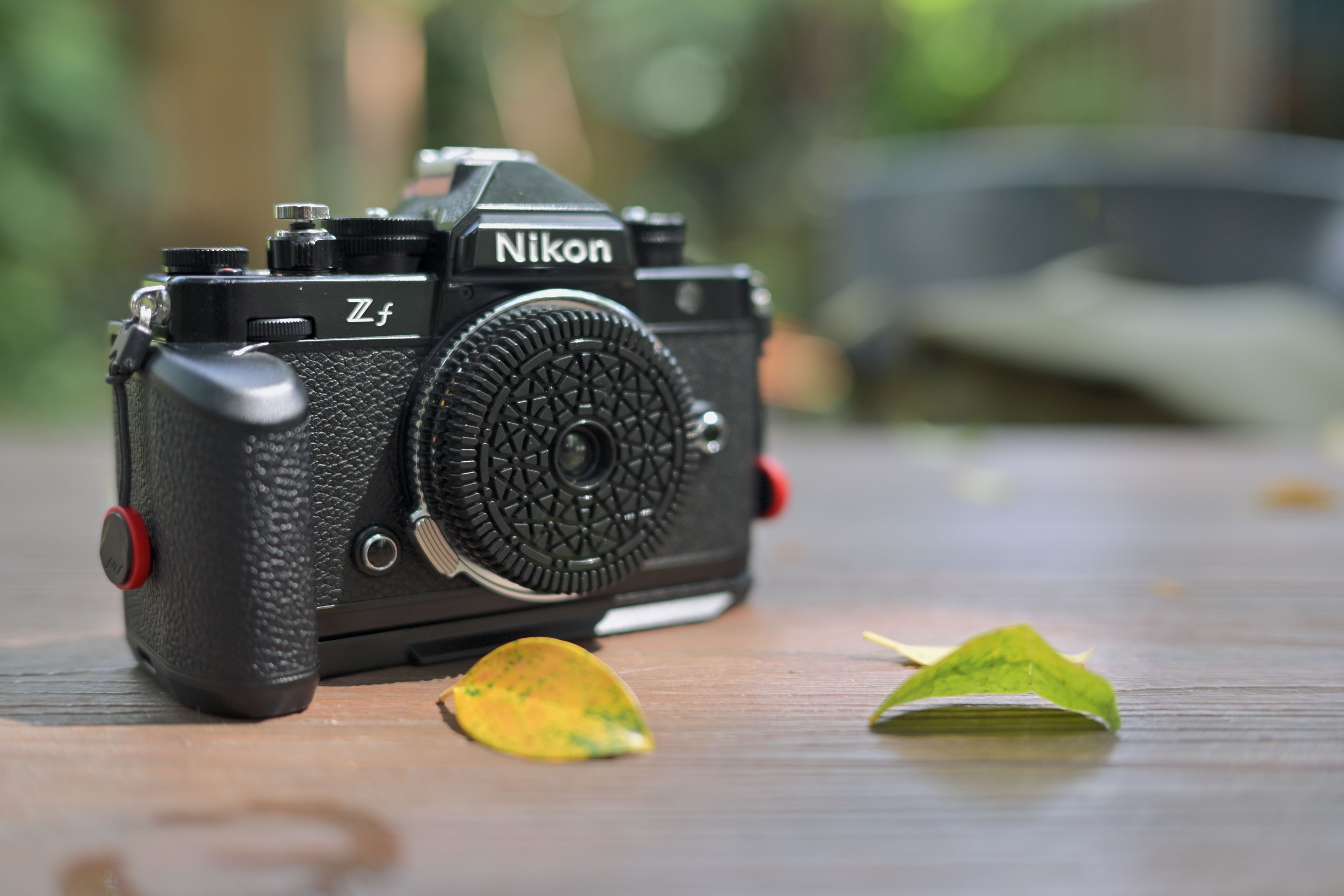 A vintage-style mirrorless camera with detailed grip on a wooden surface with scattered leaves outdoors.