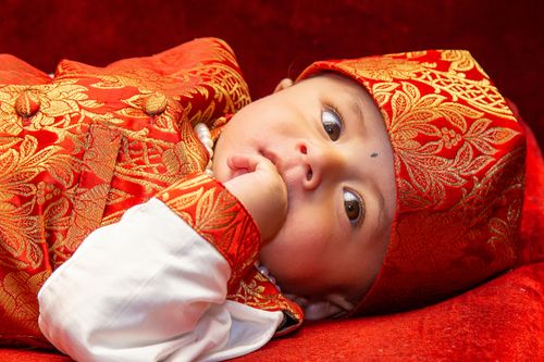 Free Charming baby in ethnic attire with gold embroidery resting peacefully on a vibrant red surface. Stock Photo