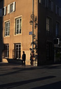 A person walks along a shadowed street corner in Nancy, France, during sunset.