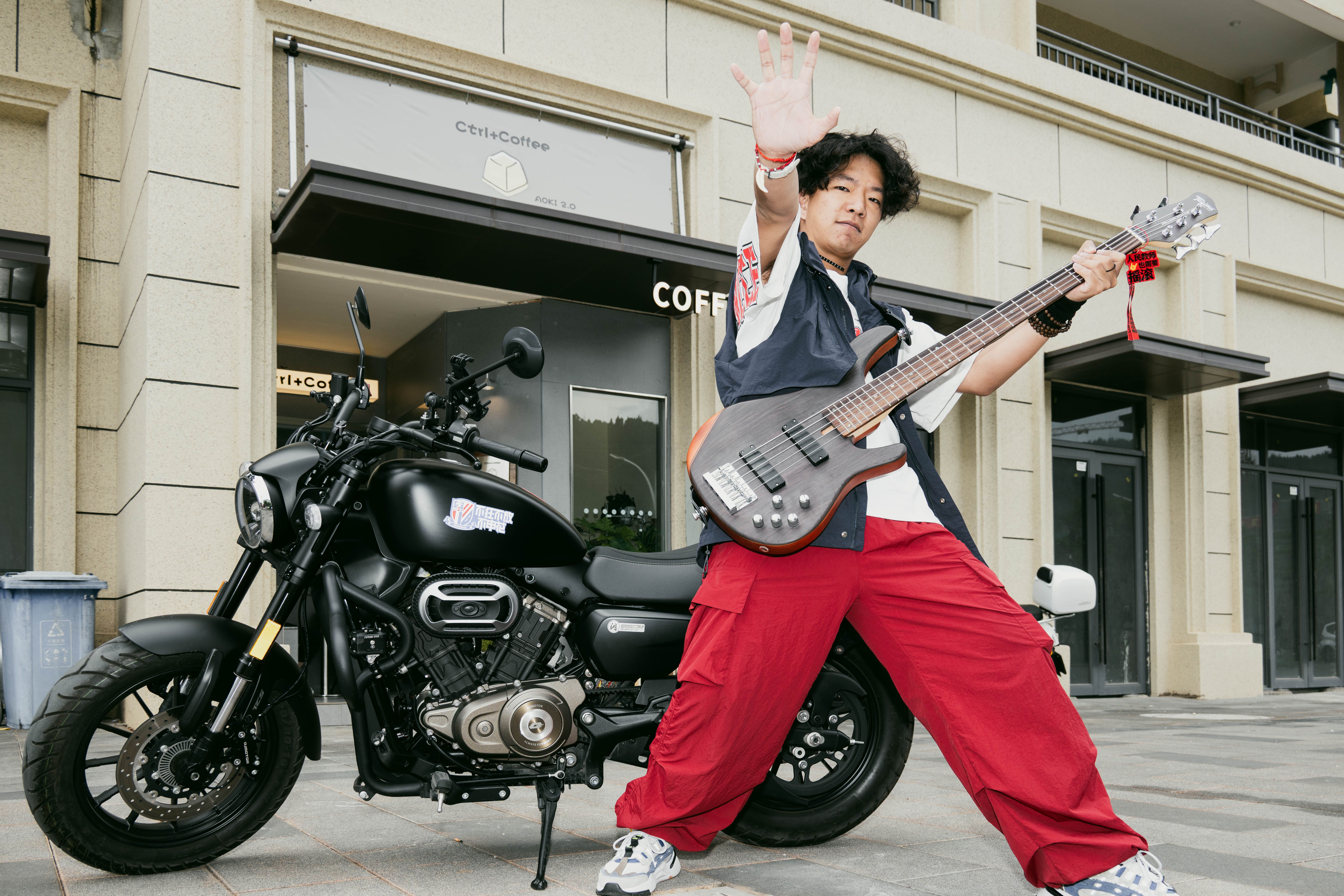 Musician holding an electric bass guitar beside a motorcycle outside a coffee shop.