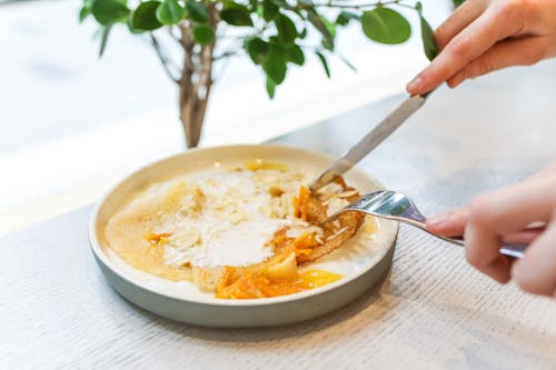 Free Unrecognizable crop person eating sweet tasty pancakes served on white plate with fork and knife while sitting at wooden table in light room Stock Photo