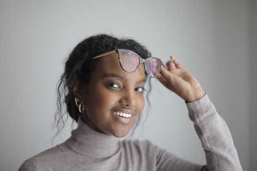 Portrait of a smiling woman in a turtleneck sweater adjusting her glasses indoors.