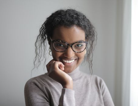 Portrait of a smiling woman with curly hair wearing glasses and a turtleneck indoors.