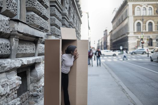 Curious African American female in casual wear hiding in own carton box on asphalt sidewalk near city road in daylight