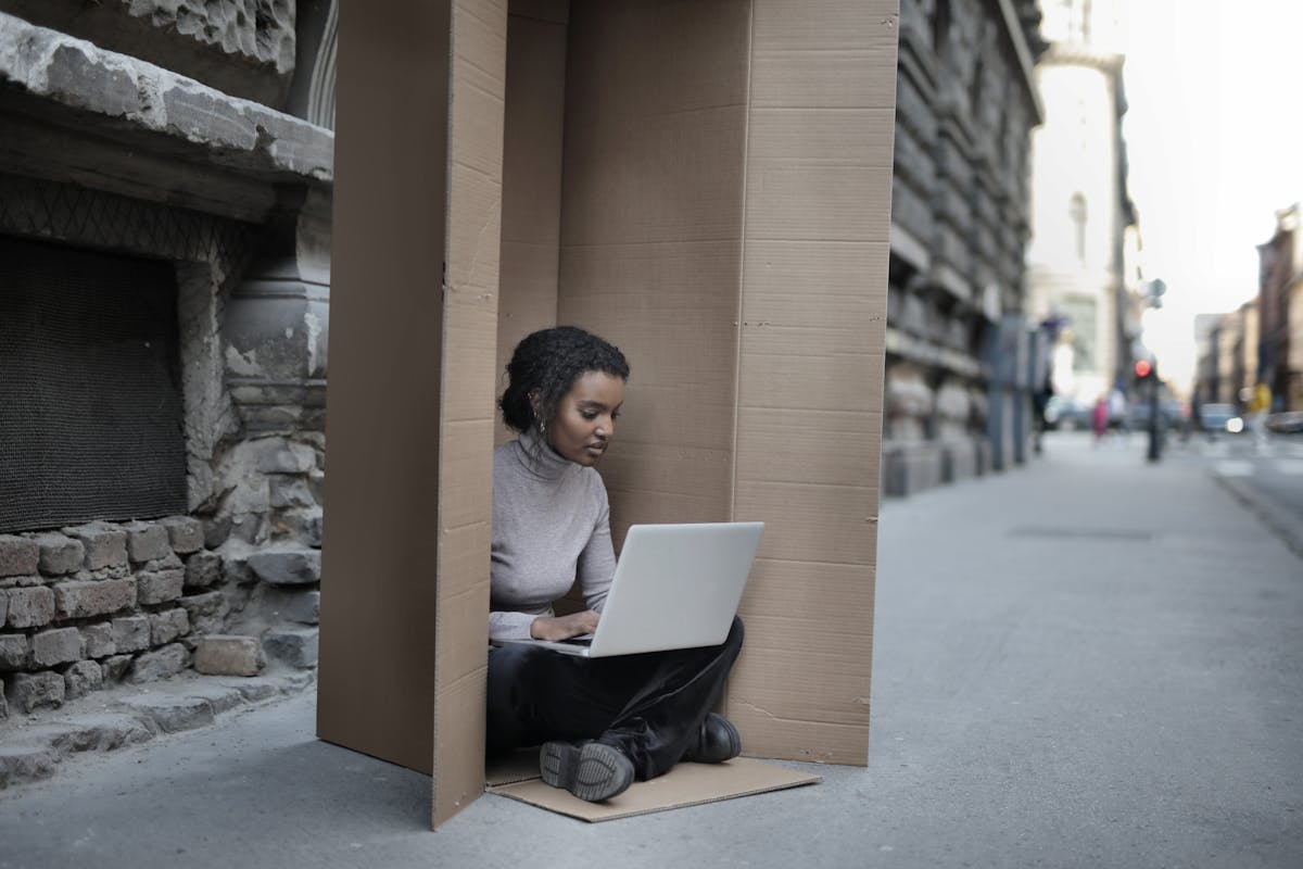 Young female introvert in casual clothes typing on netbook while sitting with crossed legs inside cardboard box located on asphalt road in city in afternoon