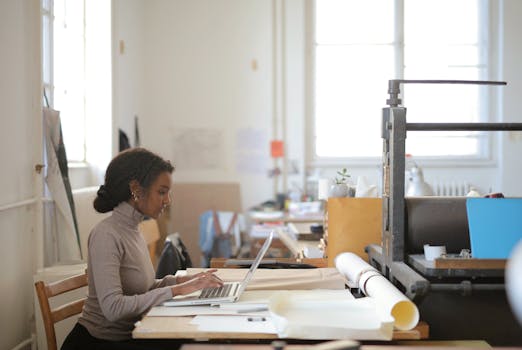 A woman engaged in remote work using a laptop in a bright, creative studio workspace.