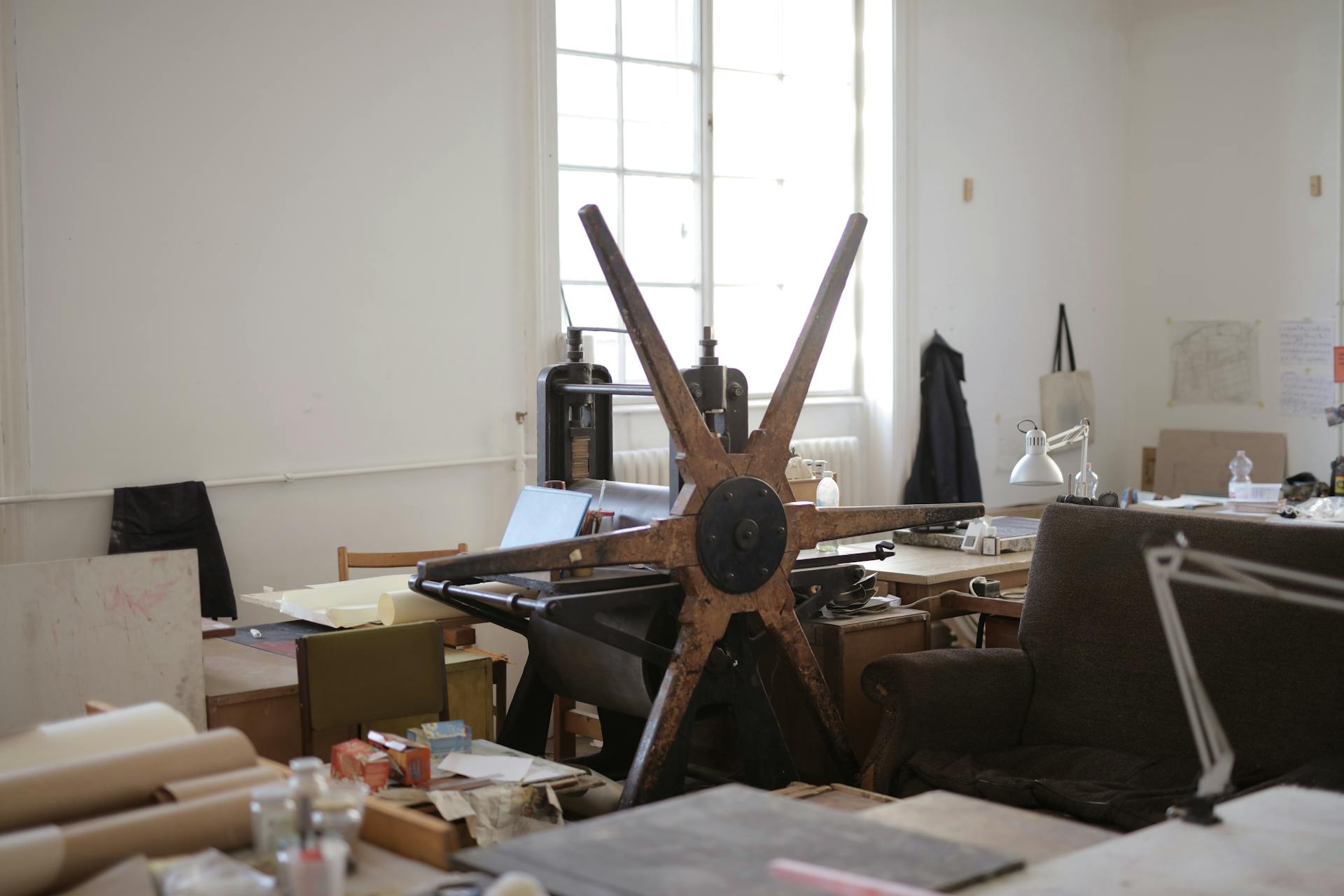 Workroom with paper rolls on table and white wall with notes and paper rolling machine with wooden mill shape wheel in afternoon