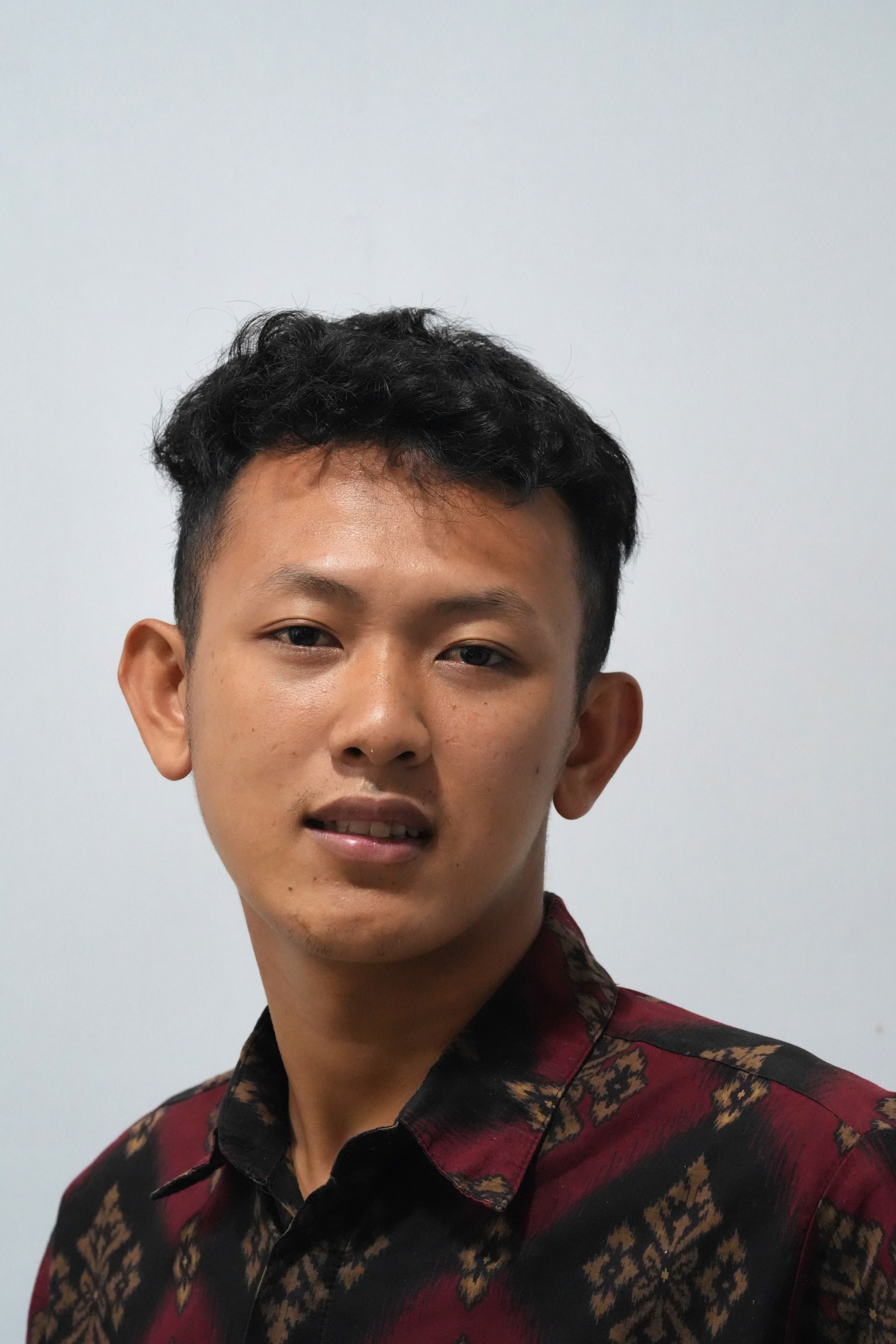 Close-up portrait of a young man wearing a patterned maroon shirt against a neutral background.