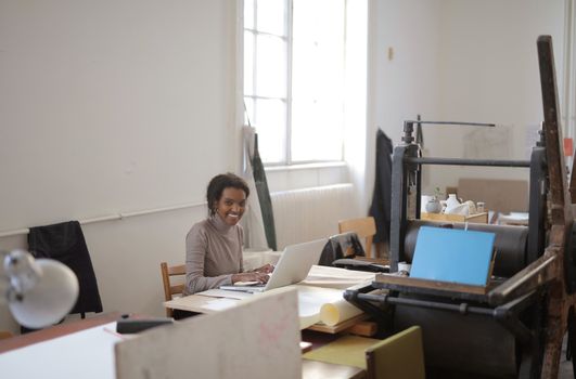 A woman smiles while working remotely on her laptop in a bright office space with a large window.