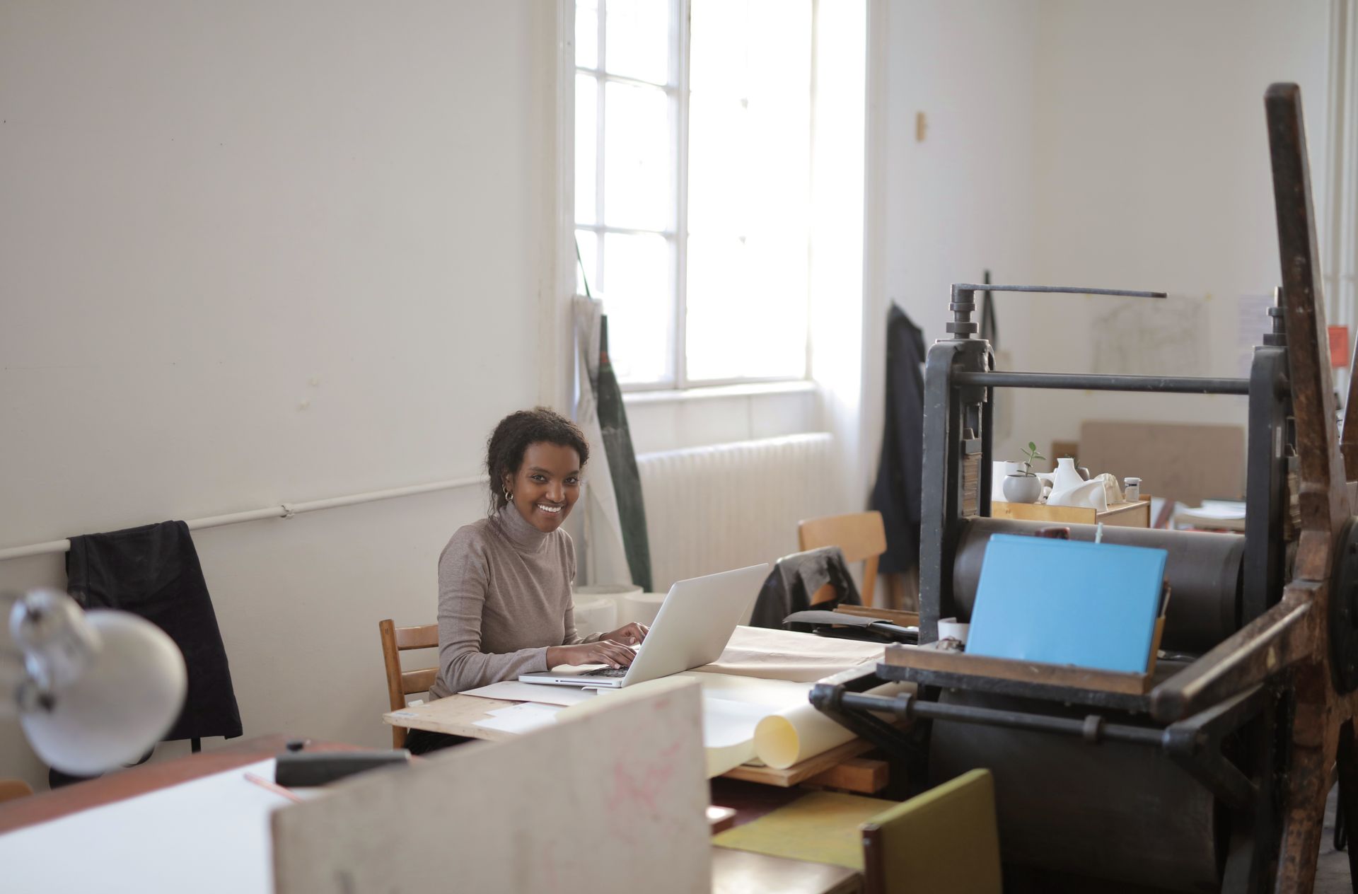 A woman smiles while working remotely on her laptop in a bright office space with a large window.