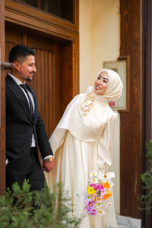 Free A joyful wedding couple holding hands outside a wooden doorway, capturing a moment of happiness. Stock Photo