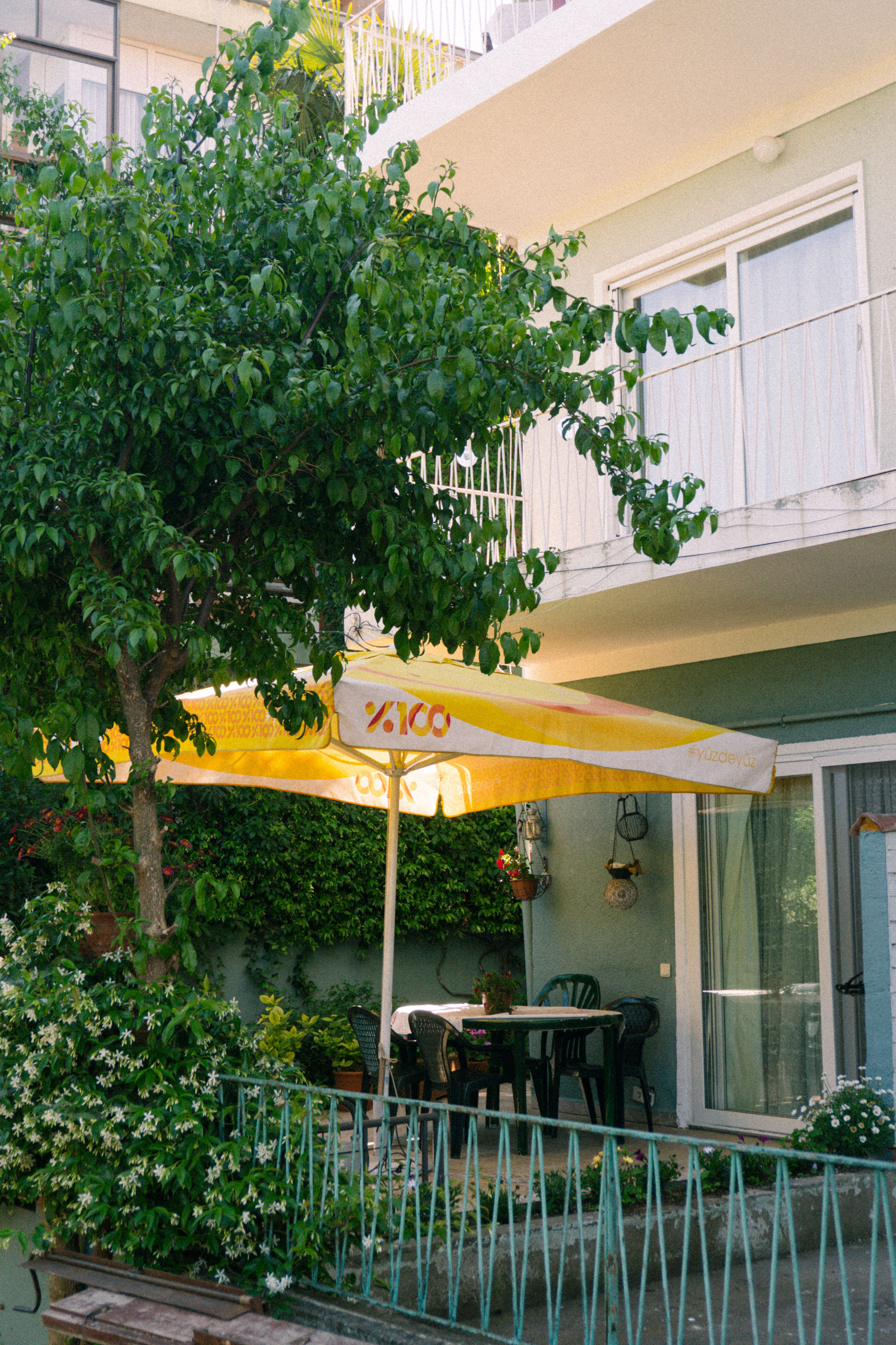 A cozy patio scene with a yellow umbrella, leafy trees, and outdoor seating in summer.
