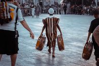 Ethnic boy wearing authentic African costume carrying big timbrels and walking on city square surrounded with people before performance
