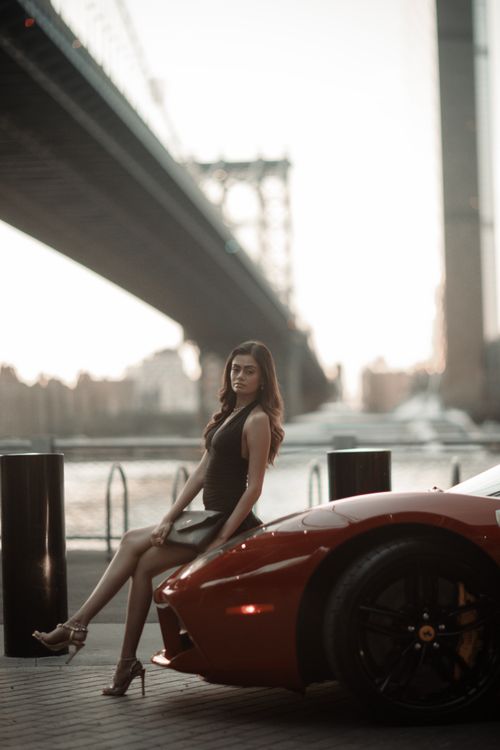 Free Chic woman in high heels poses by a red sports car near an iconic bridge. Stock Photo