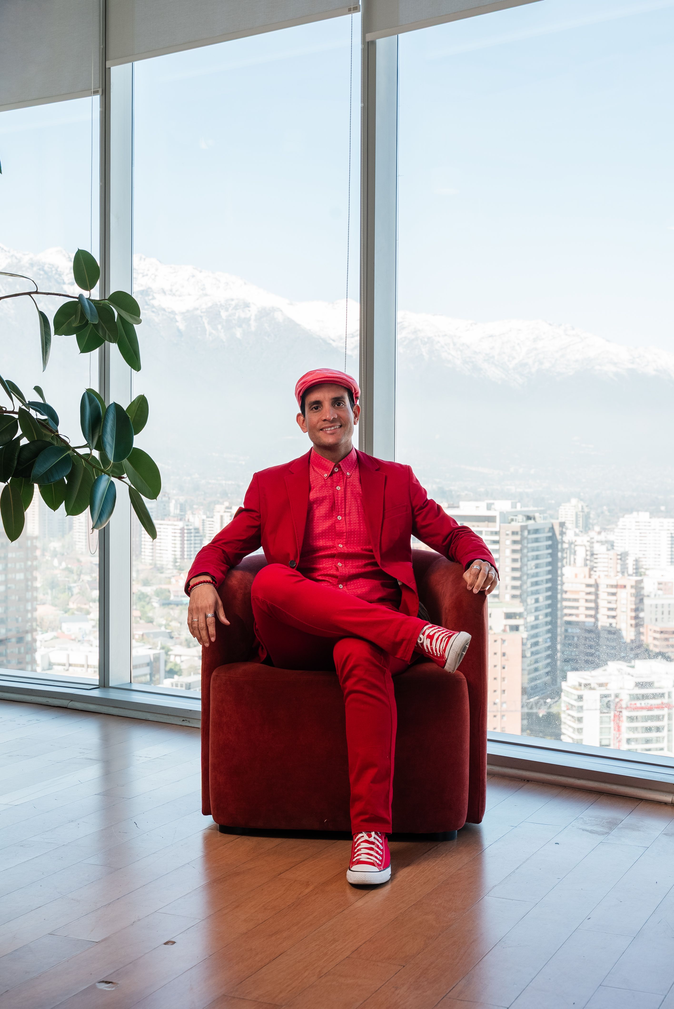 Confident man in a vibrant red outfit sits in a modern office overlooking a cityscape.