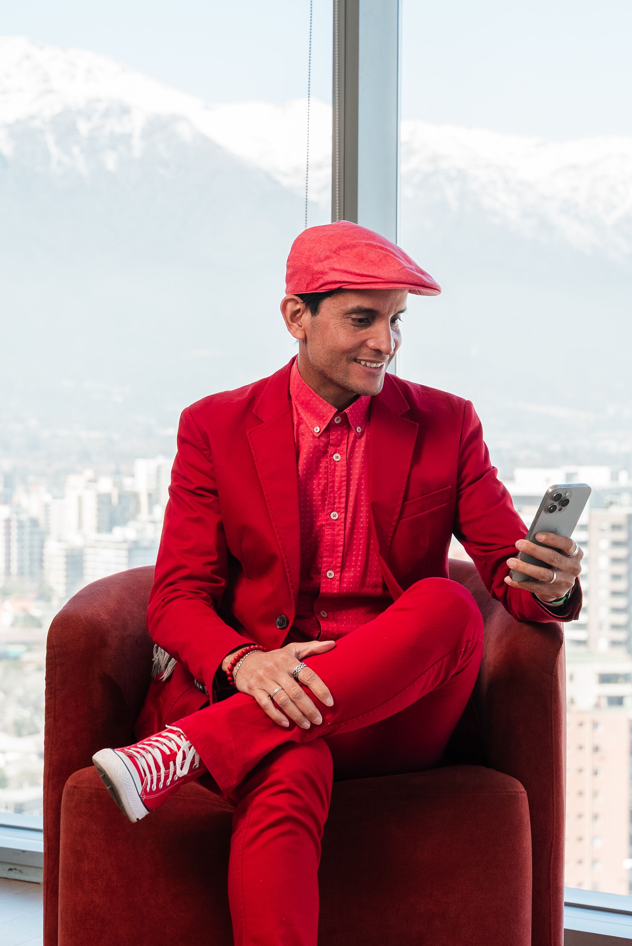 Man in red suit sitting on chair indoors using a smartphone with city skyline view.