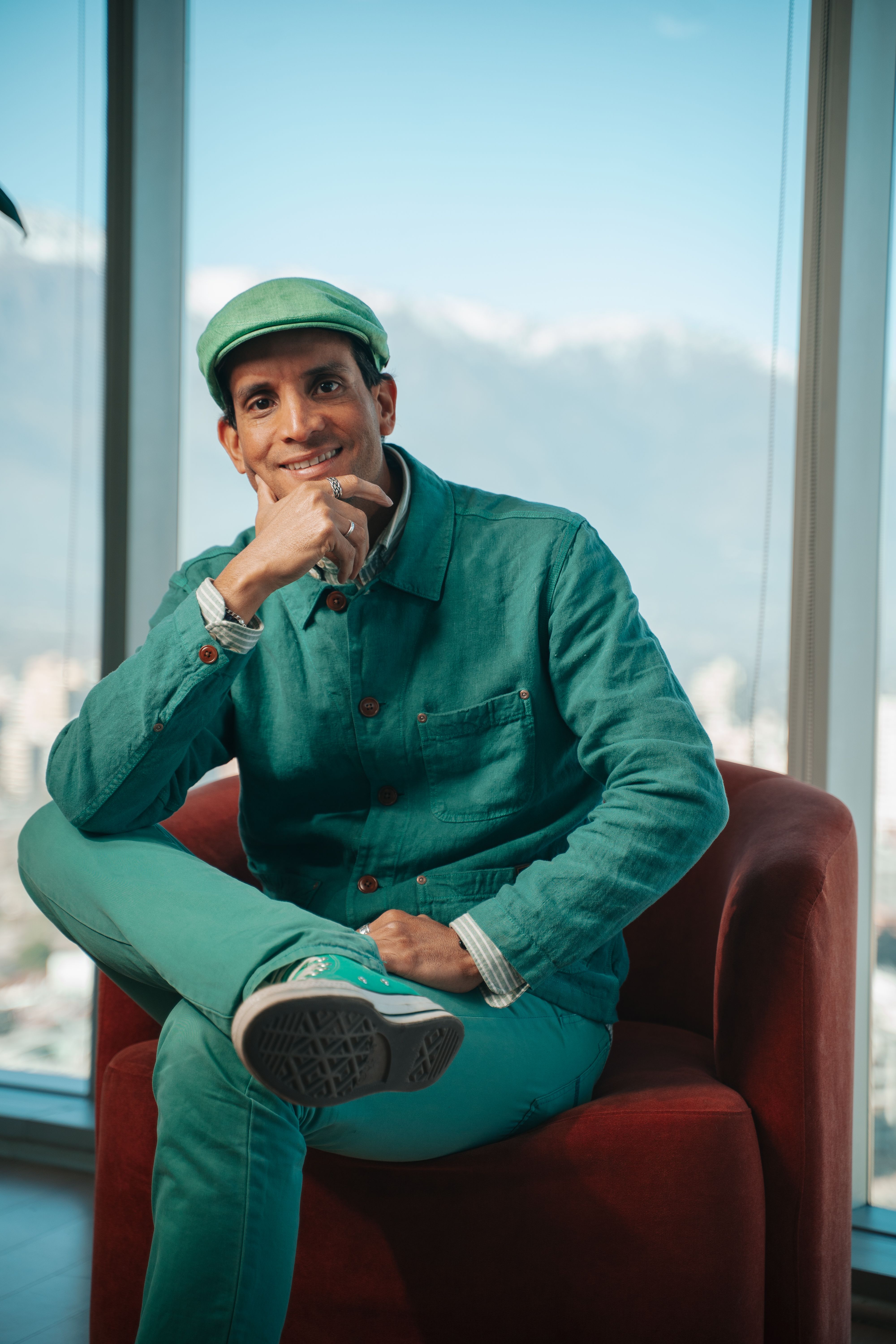Fashionable man dressed in green sitting indoors with cityscape view through large windows.