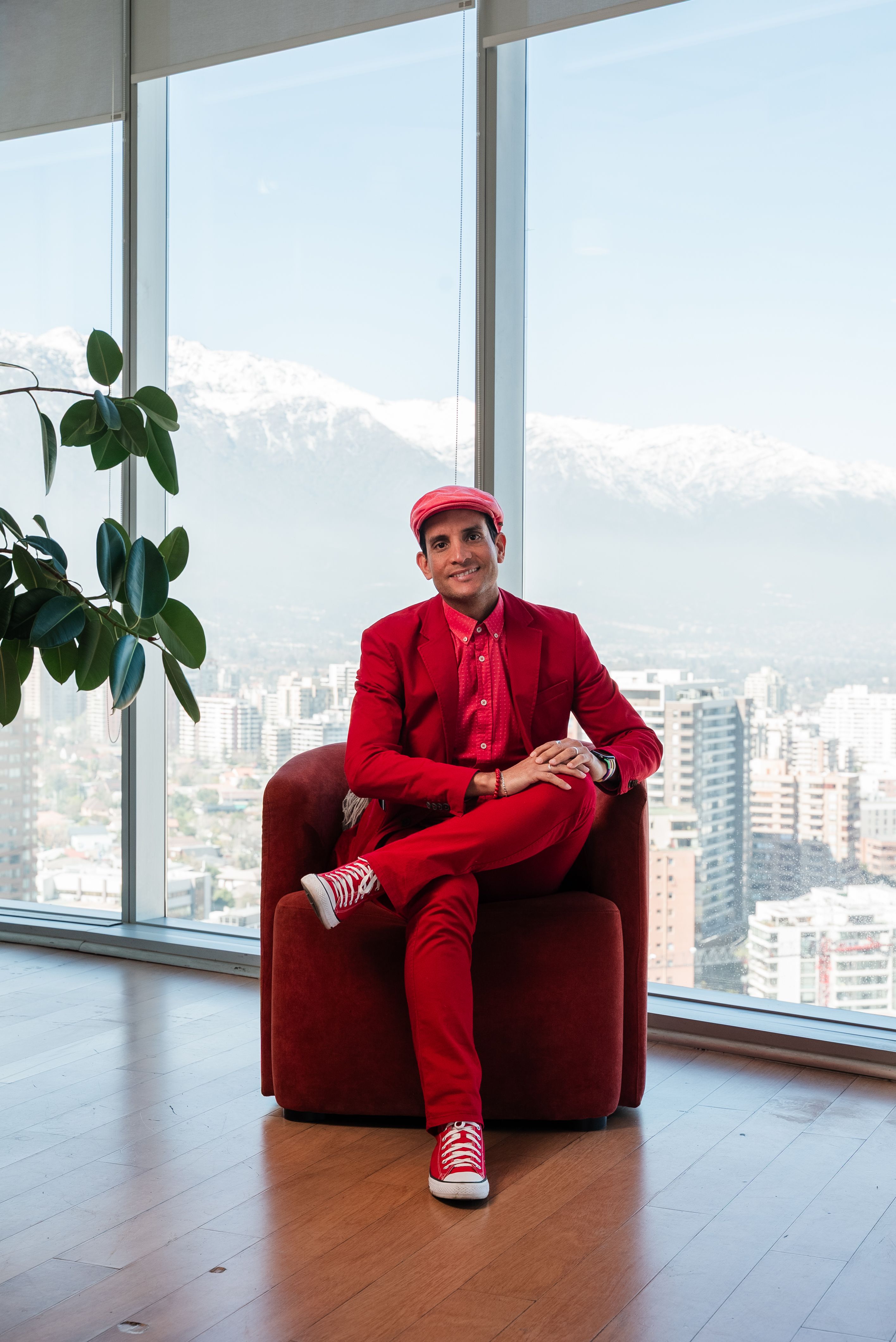 Man in red suit sitting in an office with a city skyline and mountains backdrop.