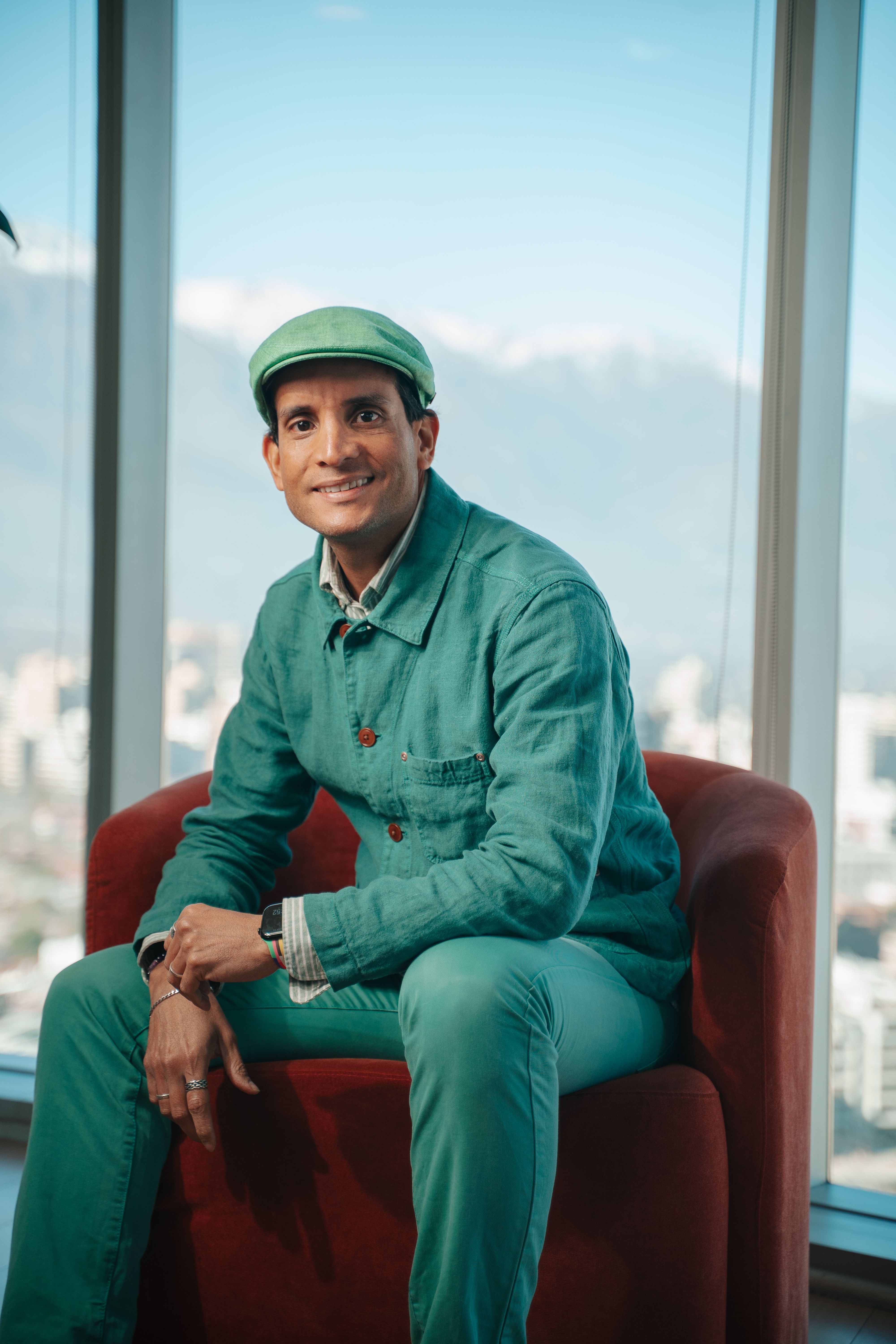A man in a green outfit sitting indoors with a cityscape background.