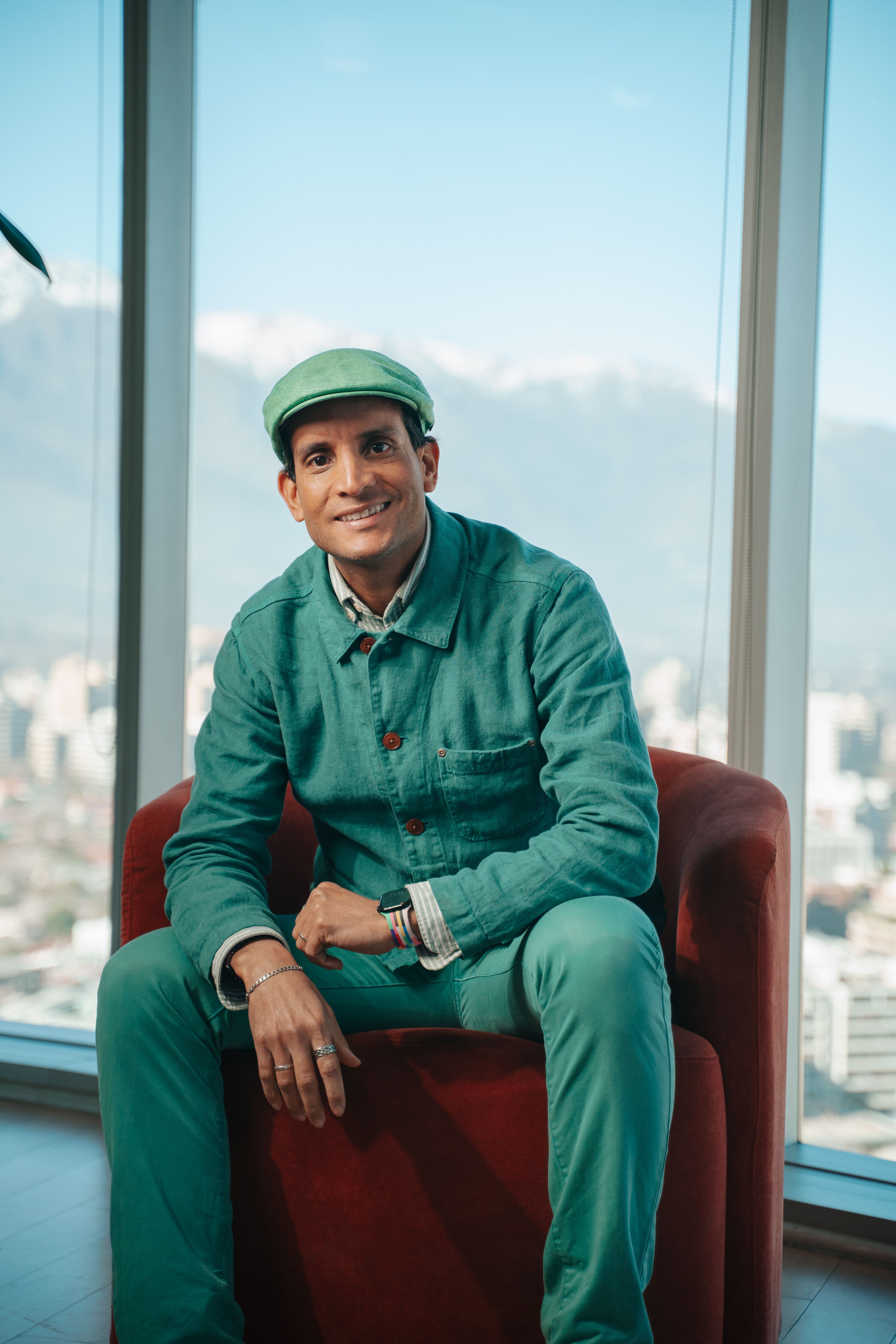 Stylish man in green attire sits by a window with a scenic city view.