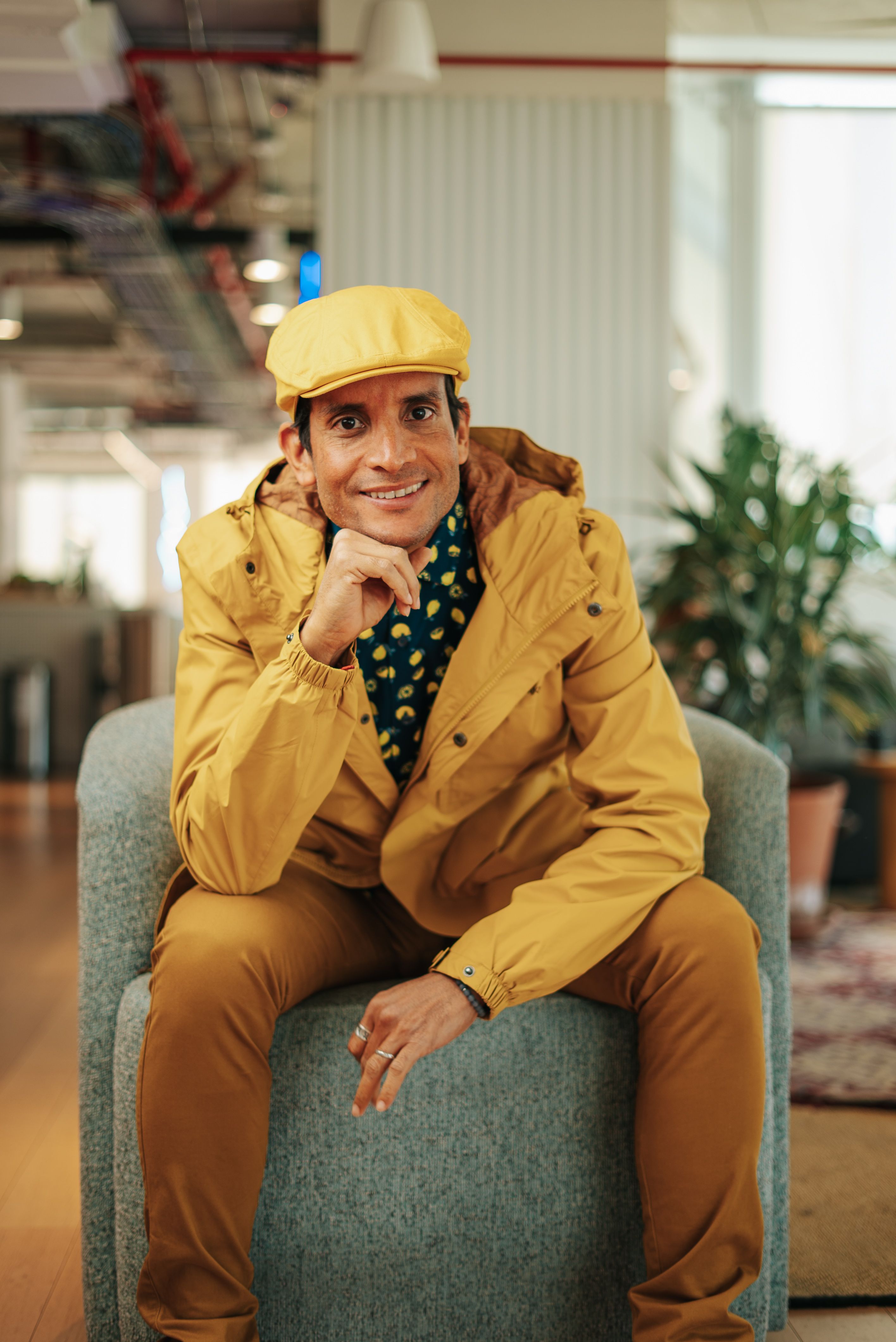Man smiling in a yellow outfit sitting on a chair in a modern indoor setting.