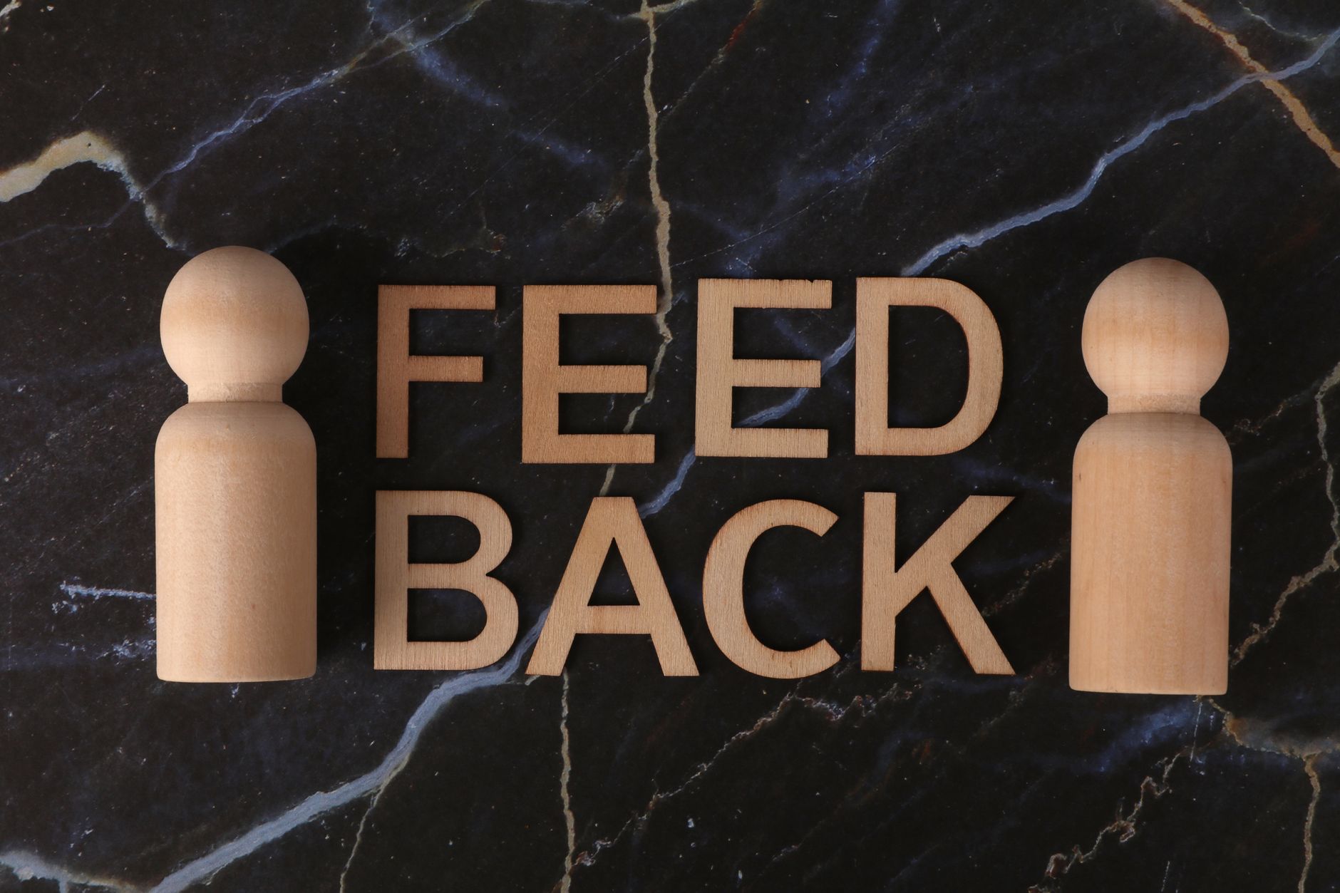 Letras de madeira formando a palavra 'Feedback' com figuras em pé sobre fundo de mármore, ilustrando a importancia dos