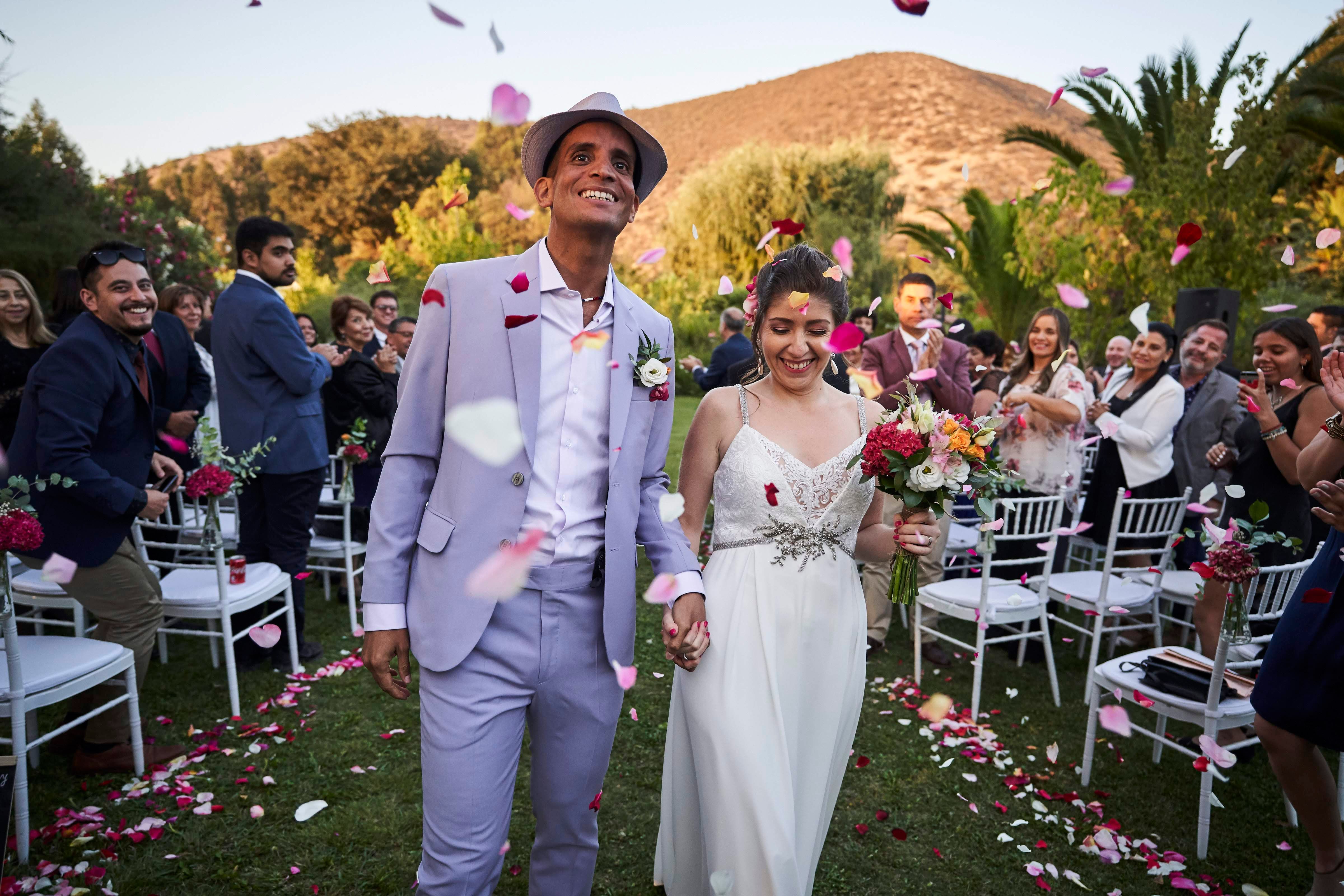 Newlyweds walk down the aisle outdoors amidst cheering guests and falling petals in Calera de Tango.
