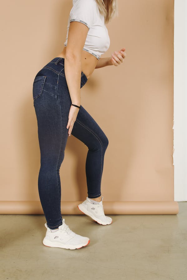 Side view of crop faceless sporty blond lady in tight dark blue jeans and white sneakers standing leaned on one leg near beige wall