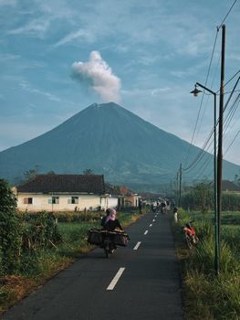 印尼一条宁静的乡村道路通往壮丽的冒烟火山，在晴朗的天空下，周围被郁郁葱葱的绿植环绕。 - 相关推荐