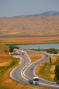 Aerial view of a winding road through picturesque hills, with trucks and cars, adjacent to a serene lake.